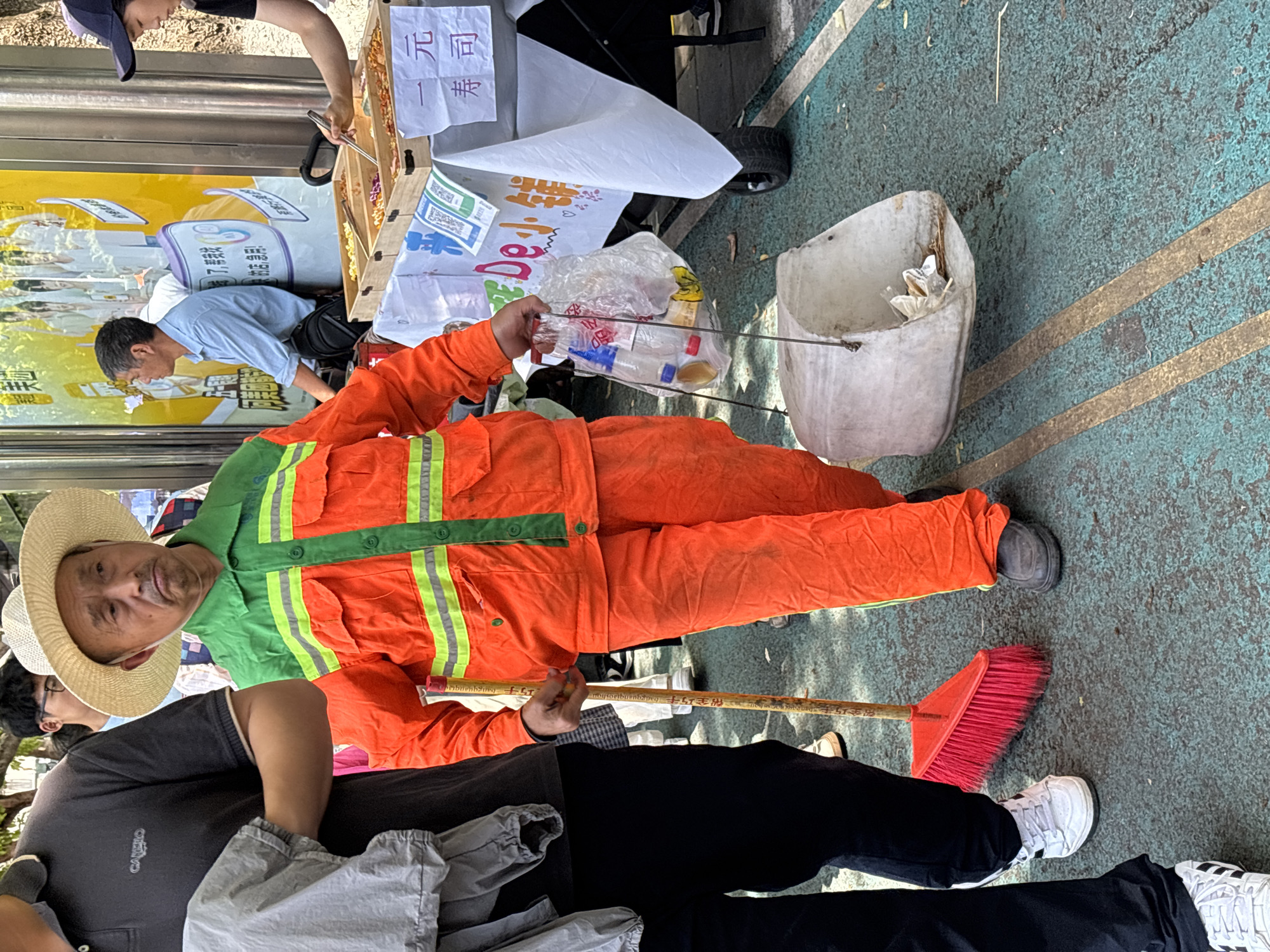 The image shows a man in an orange uniform, likely a sanitation worker, holding a red broom and a trash picker with a plastic bag of trash. He also has a straw hat on his head. Beside him, another individual is partially visible wearing a gray t-shirt and dark pants. The environment appears to be a public area with painted stripes on the ground. Behind the sanitation worker, a small cart can be seen, featuring advertisements written in Chinese. Based on the writing system and general appearance, the image could be from Jiulongpo District, China. There are a few other people in the scene in the background. One man is reaching to get an item from a small cart and another person behind them appears to be selling the items.