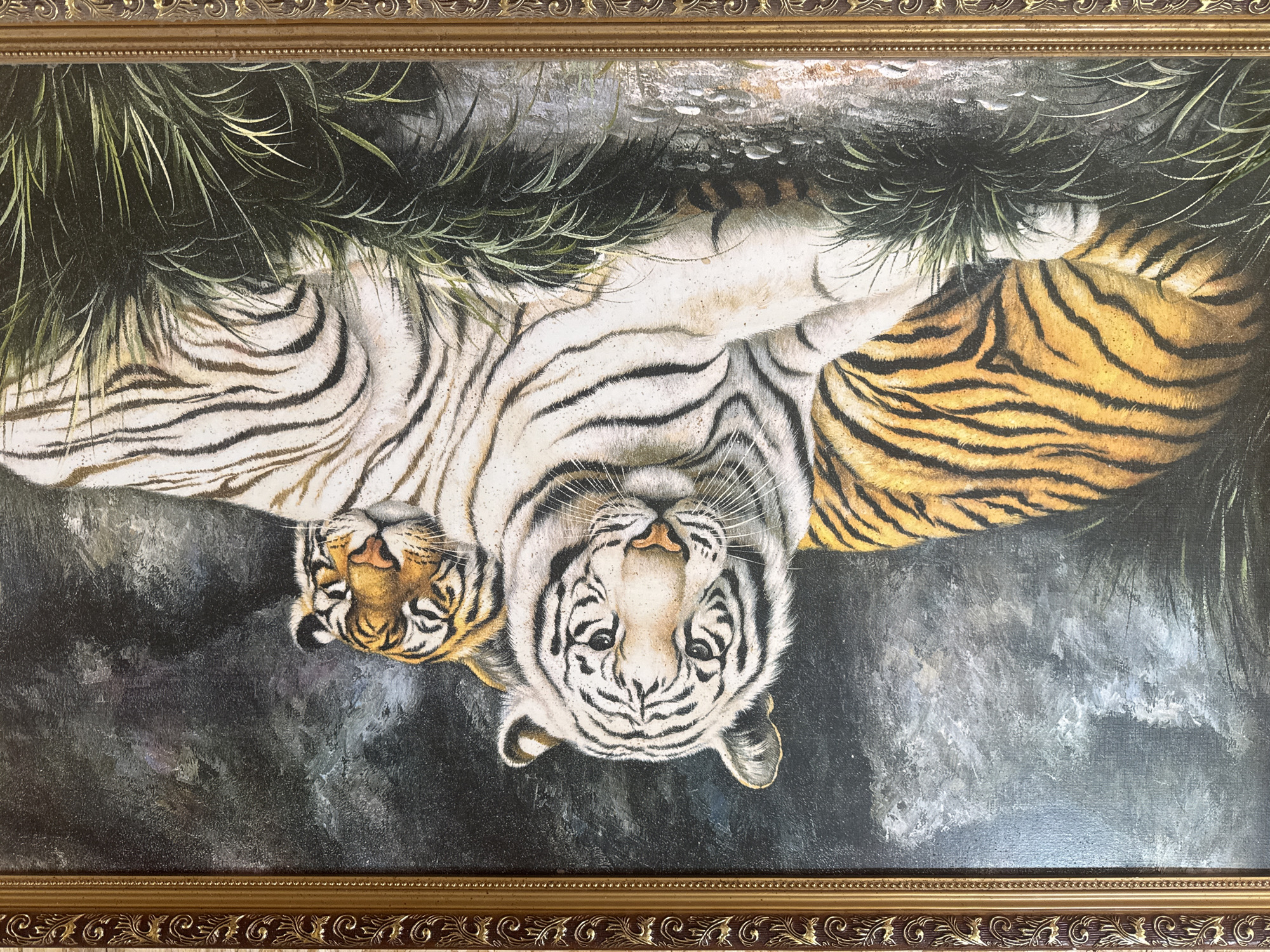 This is a framed oil painting depicting two tigers resting in tall grass. The tiger in the foreground is white with black stripes and is looking directly at the viewer. Behind it and to the right, a second tiger, with orange and black stripes, rests its head on the white tiger's back, its eyes closed. The background is dark and textured, suggesting a rocky or wooded environment. The painting is housed in an ornate, gilded wooden frame with decorative carvings. The scene is set in Protasovo, Russia.