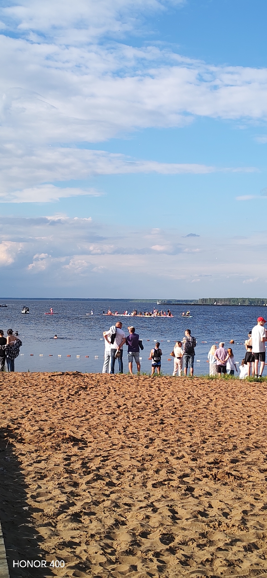 The image captures a wide shot of a body of water, likely a river or lake, under a partly cloudy sky. In the foreground, a sandy beach with tire tracks is visible, with the "HONOR 400" watermark in the bottom left corner. Along the water's edge, a line of people are gathered, some standing and conversing, others with their backs to the camera. Several individuals are in the water, swimming or standing.
Further out on the water, a large group of people are on paddleboards or rafts, forming a procession. Smaller boats and a distant shoreline with trees complete the scene. The overall atmosphere appears to be a recreational event or gathering by the water.
While the image does not explicitly state its location, the presence of "HONOR 400" on a device used to capture the image could suggest a connection to technology or a specific product, but provides no geographical information. This scene is typical of many waterfront recreational areas. Given that it is a general scene of a river or lake with people, it could be any number of locations.