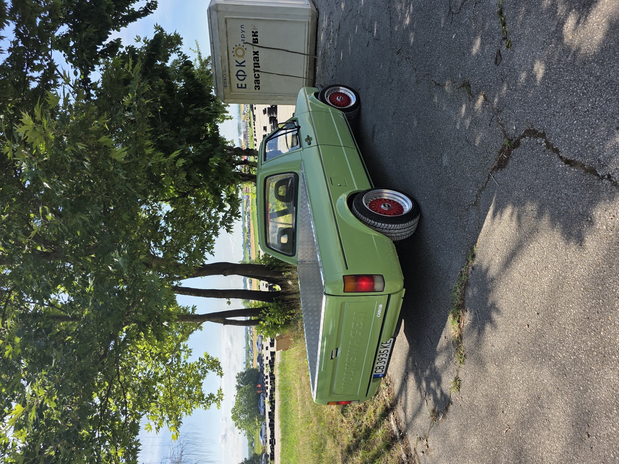 A light green 1980s Volkswagen Caddy pickup truck is parked on an asphalt surface next to a white portable building with Cyrillic text. The truck has a silver diamond plate bed liner and red-accented alloy wheels. Its license plate reads "CB 3935 KE". The portable building has text that appears to say "ЕФКО груп" and "Застраховки". The background shows trees with lush green leaves, a clear blue sky with scattered clouds, and a distant view of a race track with other vehicles and barriers. The scene appears to be an outdoor location, possibly a parking area or event venue, consistent with Akatsievo, Bulgaria, a location known for its karting track.