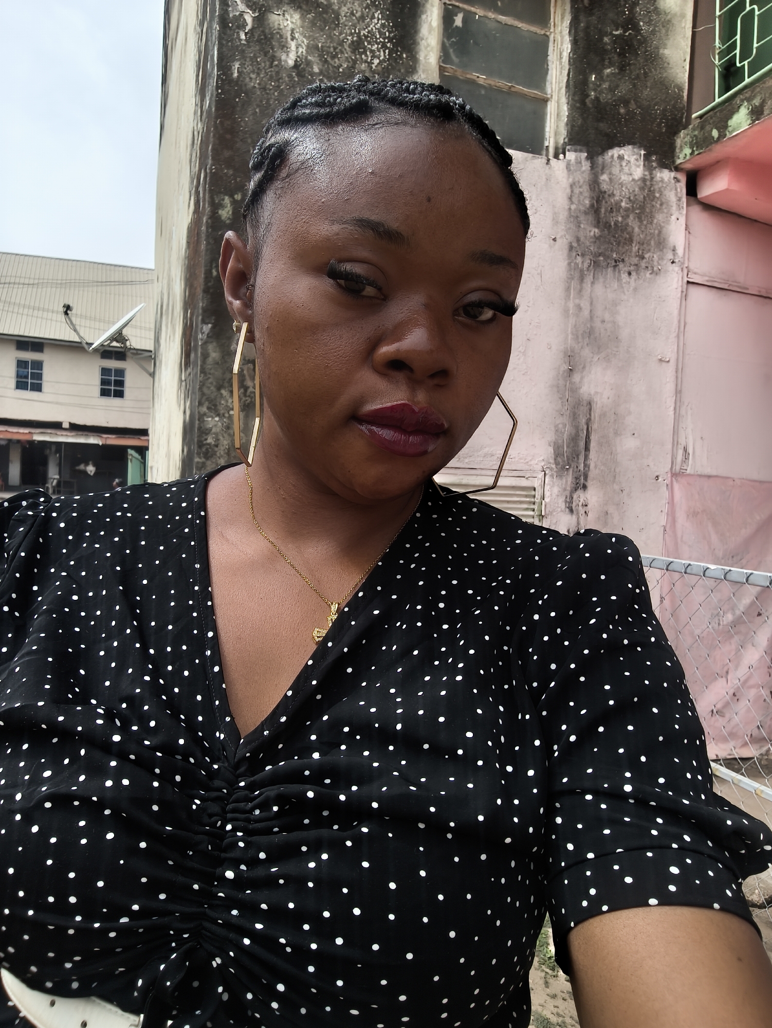 A close-up, slightly high-angle portrait captures a Black woman from the chest up, likely a selfie. She has braided hair, pulled back, and features dark eyes with long false eyelashes. Her full lips are adorned with dark plum or burgundy lipstick. She wears large, gold-colored hexagonal hoop earrings and a delicate gold chain necklace with a small pendant, possibly a cross. Her attire is a black top or dress with white polka dots, featuring a V-neckline and gathered, ruched fabric at the bust. Her expression is neutral and serious as she looks directly at the camera.
The background reveals an urban setting under an overcast sky. To the left, a light-colored, two-story building with multiple windows and a white satellite dish on its roof is visible. To the right, a tall, weathered, light-toned wall, stained with dirt or mildew, dominates the midground. Behind this wall, parts of another building, possibly pink or red, with green lattice work and a chain-link fence, suggest a somewhat aged or neglected environment typical of urban areas in places like Jos, Nigeria. No visible text is present in the image.