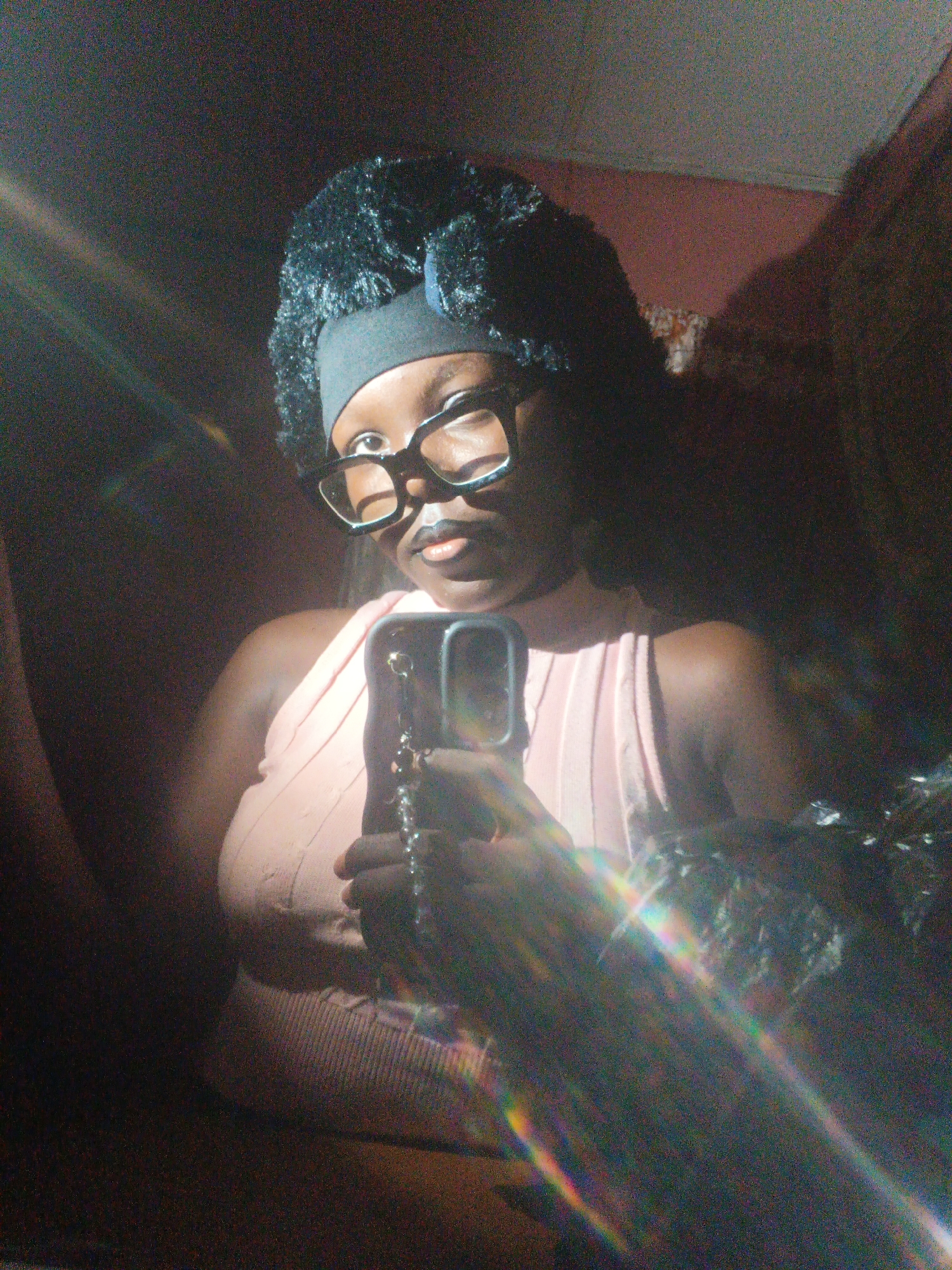 A person with dark skin, wearing black-rimmed glasses and a black headband with a furry black hat, is taking a selfie in a mirror. They are wearing a light pink, sleeveless top. Their lips are painted with dark lipstick. The background is dimly lit, with a hint of a pink wall and some shadows. There are lens flares across the image, creating streaks of light and color. The person is holding a cell phone in their right hand, and a chain attached to the phone is visible. The overall lighting is poor, and the image is grainy.