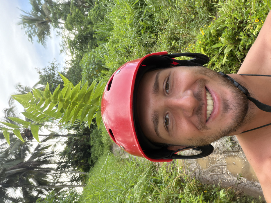 A selfie of a smiling man with a reddish helmet, set against a backdrop of lush, green vegetation including a prominent fern seemingly growing from his helmet. Palm trees and other foliage reach towards a slightly overcast sky. The scene suggests a tropical location, possibly a rainforest or jungle, with muddy ground visible. The man's casual attire and the helmet hint at an outdoor adventure activity.