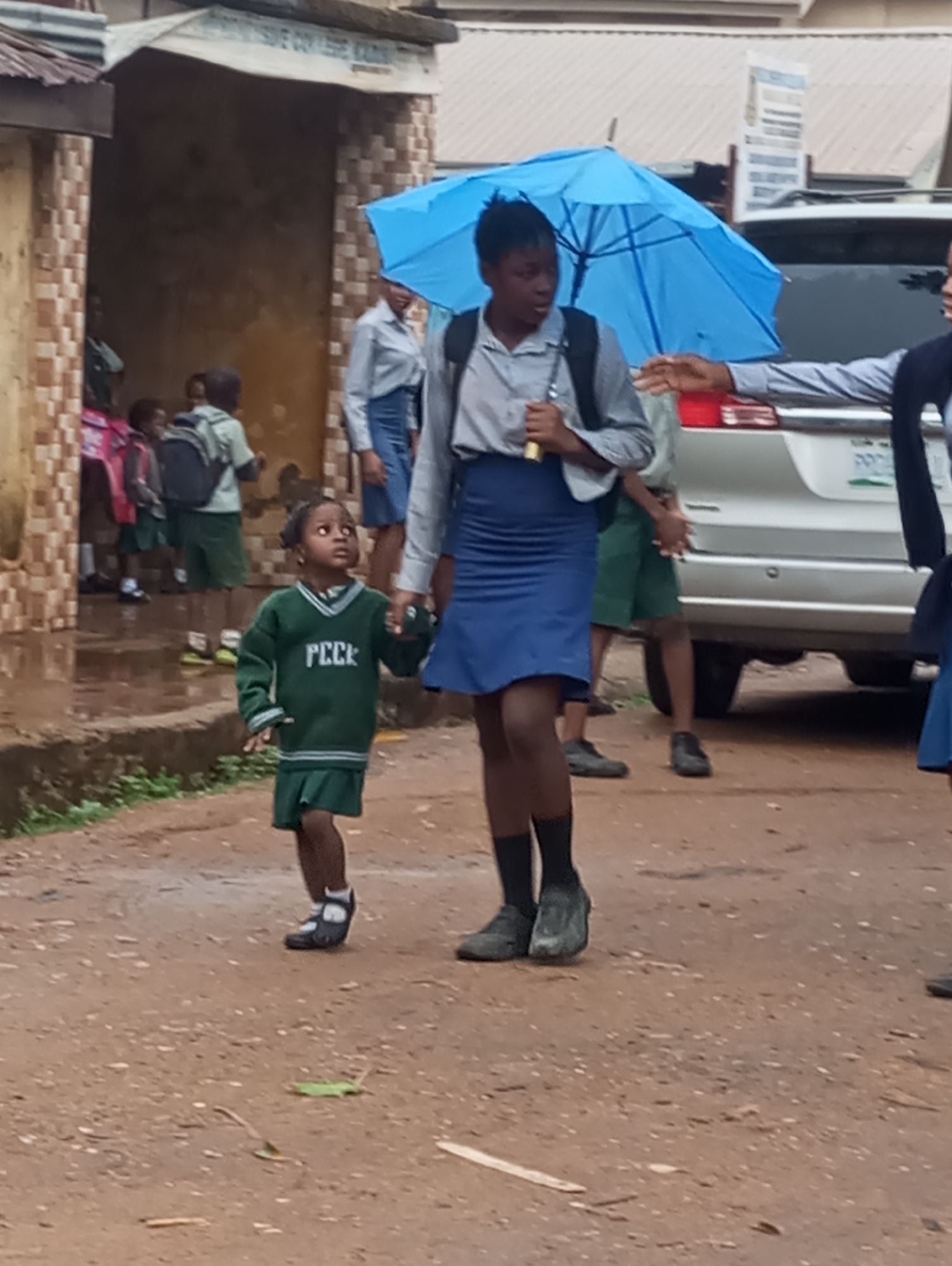 In Sabon Tasha, Nigeria, a young girl in a green school uniform is holding hands with an older girl carrying a blue umbrella. Both are walking on a dirt path. Other children in school uniforms are visible in the background, near a building and a parked car. The scene appears to be an outdoor area adjacent to a building with writing on it.