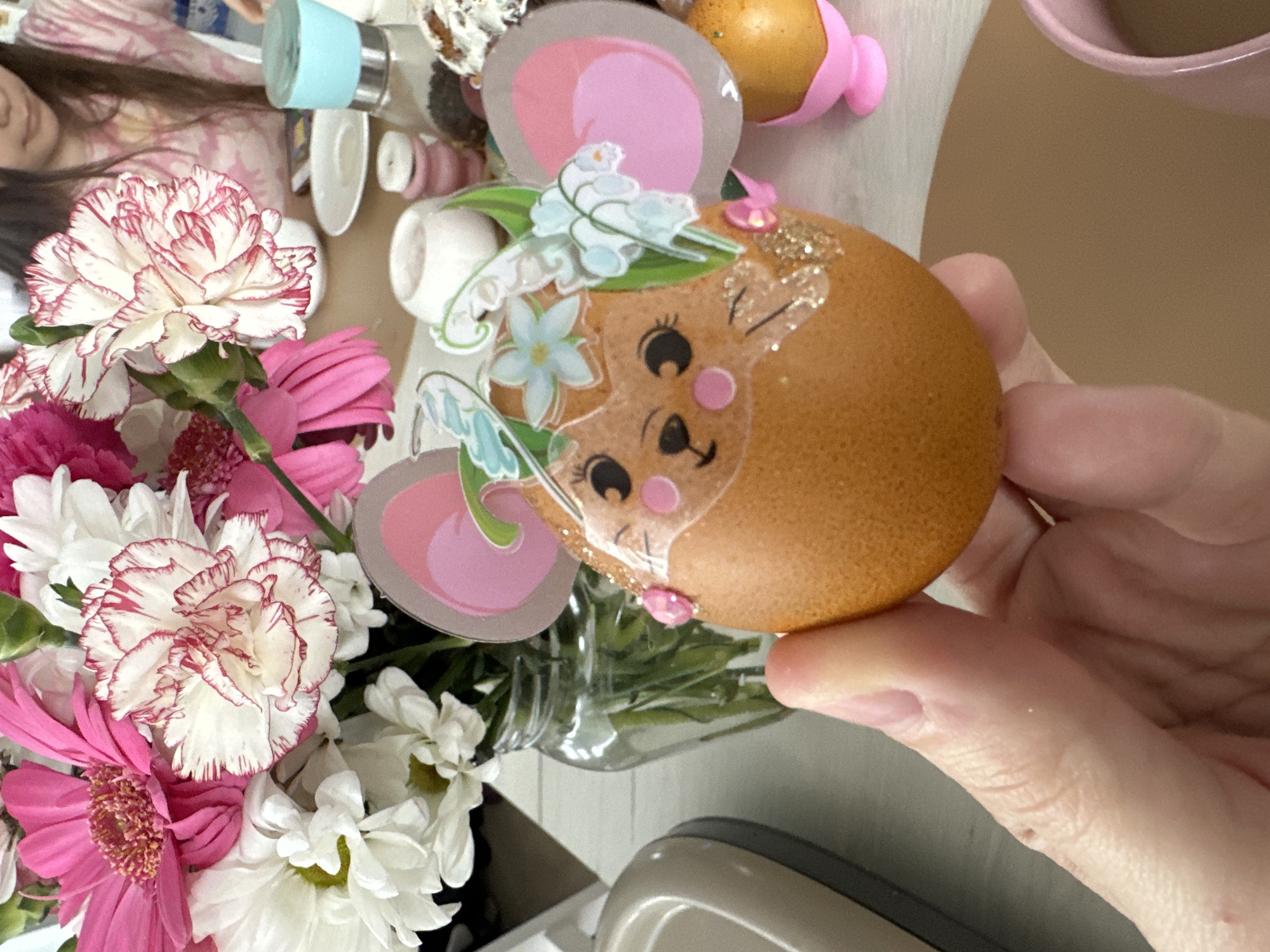 A close-up, angled shot shows a decorated brown egg being held in a person's hand. The egg is decorated with a cartoon face of a creature with pink cheek circles and glitter accents. Paper cutouts of ears and flowers are attached to the egg. Next to the hand holding the egg is a vase of pink, white, and red flowers. Other objects are scattered in the background including a teal container, white saucers, and a pink container.
