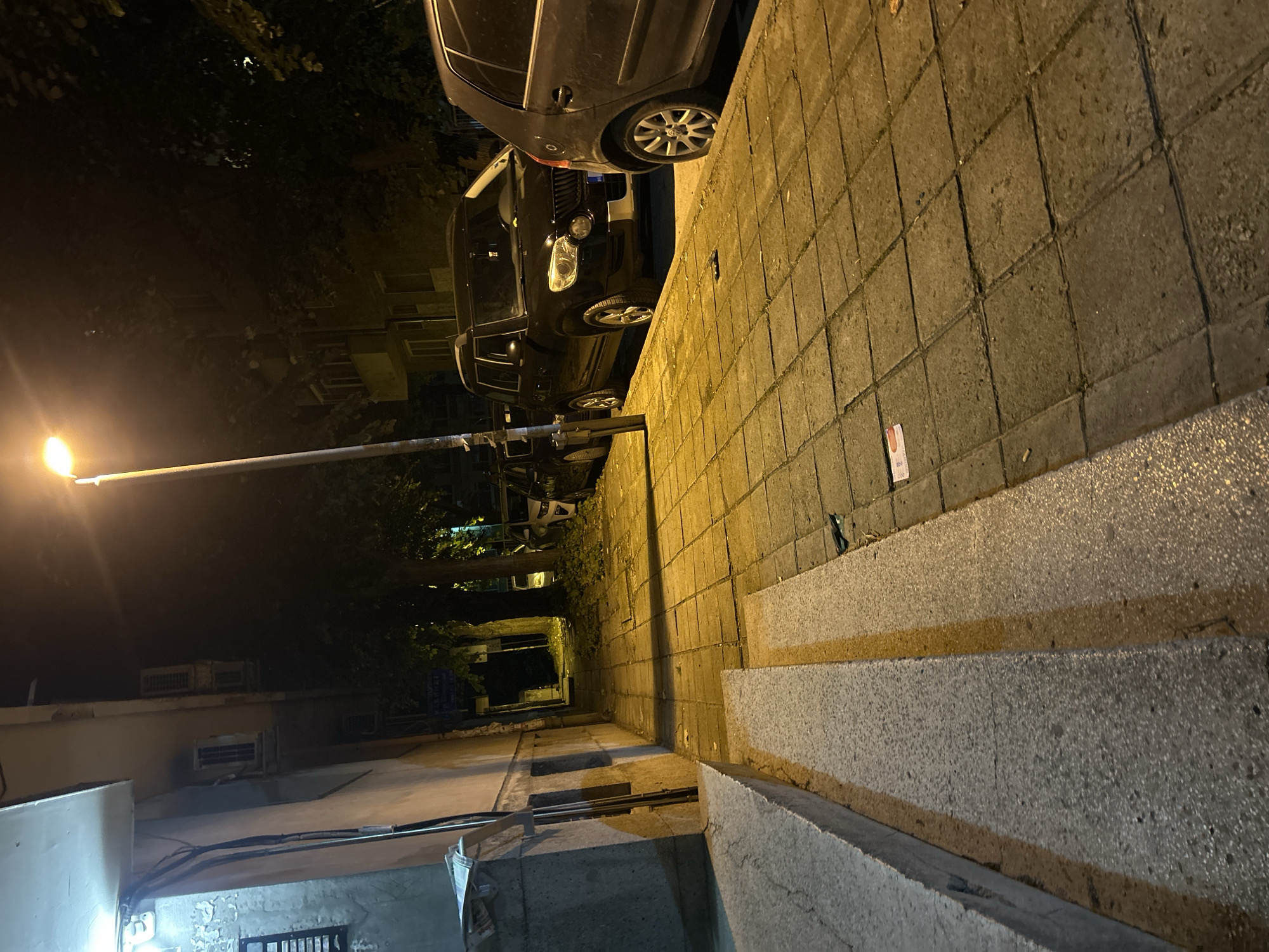 The image depicts a nighttime scene on a city street in Varna, Bulgaria. A sidewalk paved with square tiles runs alongside a building on the left and a street with parked cars on the right. A lamppost illuminates the scene with warm, yellow light, casting long shadows. Several cars are parked along the street, including a black SUV and a dark-colored sedan. The building appears to be residential, with windows and air conditioning units visible. Trees line the street, their leaves appearing dark against the night sky. The sidewalk is littered with some debris, including what appears to be a small piece of paper and a cigarette butt. The overall impression is one of a quiet, ordinary urban night.