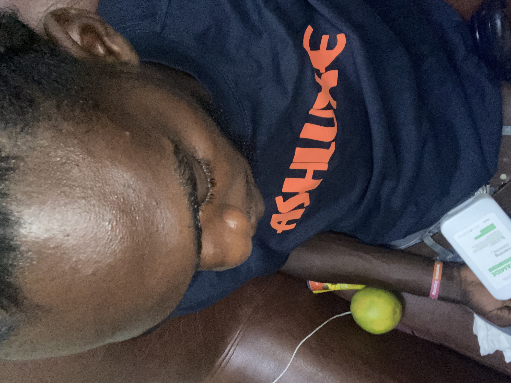 The image displays a male individual with dark skin, viewed from an elevated perspective, seated on a brown, upholstered sofa or chair. The individual's head is tilted downward, exposing a forehead with a visible sheen, and dark, braided hair. A short, trimmed beard is present on the chin and jawline. The person is wearing a dark blue t-shirt with the orange word "ASHLLUXE" printed across the chest. A pink and orange wristband, partially legible as "Reflecting Tomorrow," adorns the left wrist. The individual's left hand grasps a white rectangular object, possibly an electronic device or compact container, featuring a green label with the partially visible text "FAMIDE." Positioned on the brown seating surface next to the arm are a spherical green citrus fruit, resembling a lime or small orange, and a small crumpled white tissue. A thin white cable is also visible extending from the left side of the frame. The specific location within Lagos, Nigeria, cannot be determined from the visual evidence.