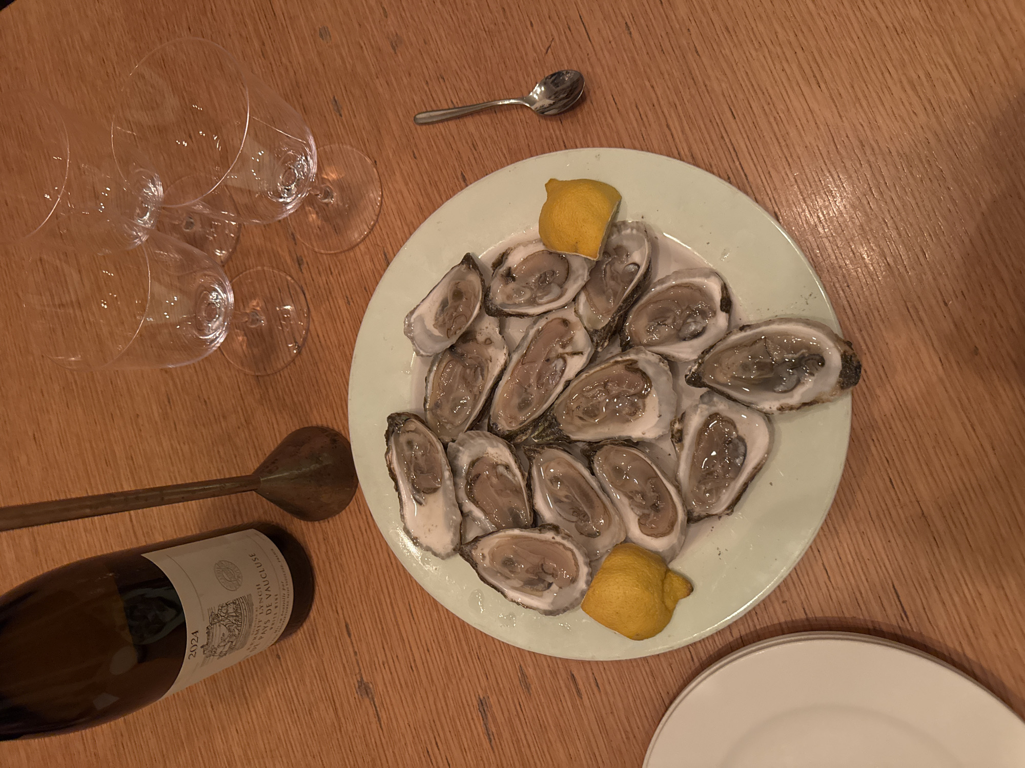 This image depicts a tabletop scene set in the city of New York, United States of America. Centrally positioned is a light green oval plate holding 15 freshly shucked oysters on the half-shell, garnished with two yellow lemon wedges. To the upper left, a dark glass wine bottle, displaying a label with "2024," "Kermit Lynch," "Pays de Vaucluse," and "White Wine," rests on its side. Three empty clear wine glasses are arranged in the upper right quadrant of the image. A small, brass-colored object with a slender handle and a wider, bell-shaped base stands upright near the wine bottle. A small silver spoon lies to the right of the oyster plate. A partial view of a white plate with a grey rim is visible in the bottom-left corner. All items are arranged on a light brown wooden table with a distinct grain pattern. The scene is illuminated by warm, indoor lighting. No individuals are present, and no actions or interactions are depicted.
