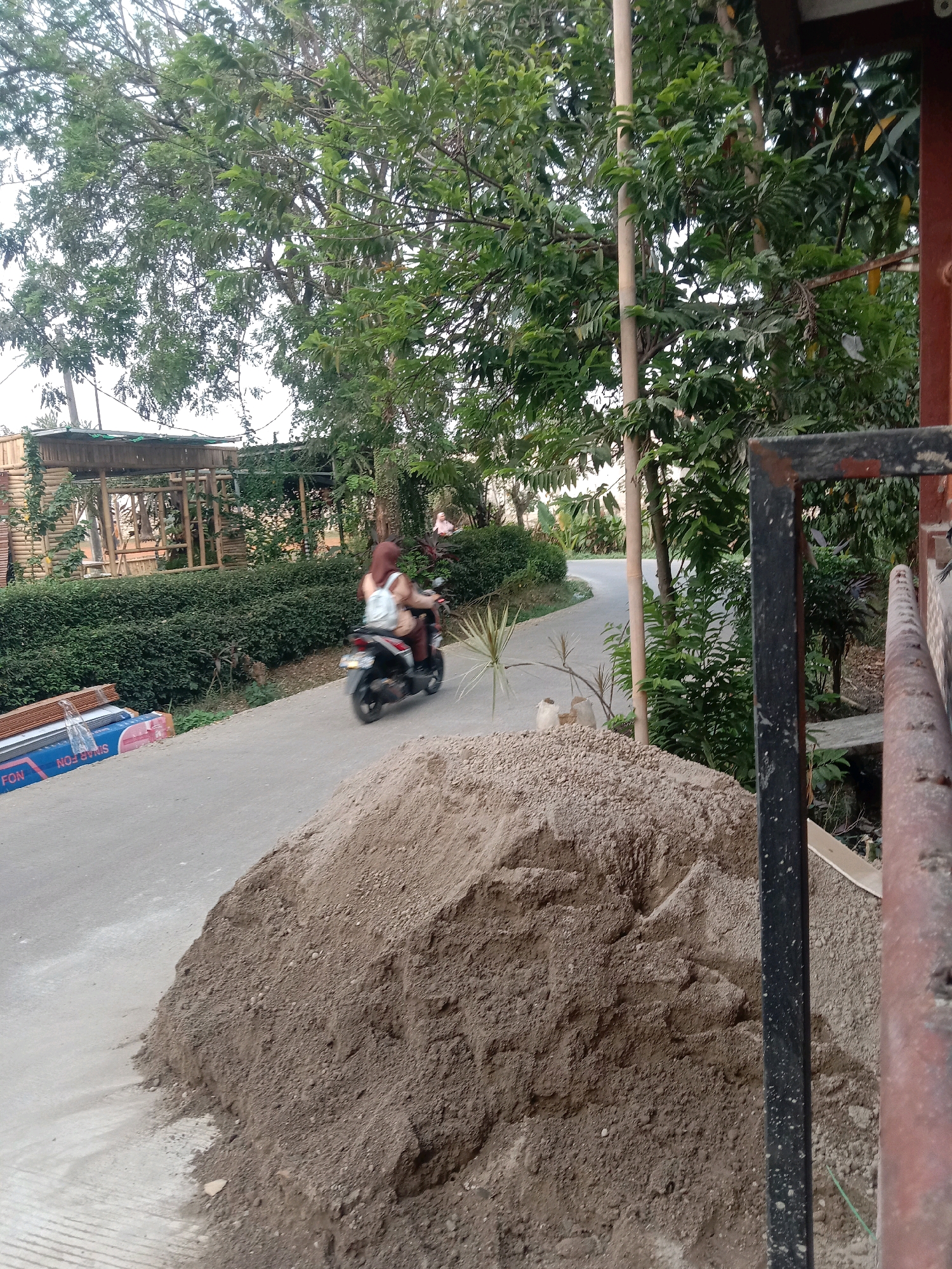 In Tajurhalang, Indonesia, a person wearing a hijab and a light-colored backpack rides a scooter down a paved road. The road is lined with lush green bushes and trees. To the left of the road, there are stacks of building materials, including what appear to be metal sheets with blue and red markings. A large pile of dirt or sand is situated on the right side of the road, close to the foreground. In the background, there is a rustic-looking bamboo structure. The lighting suggests it is late afternoon or early evening.
