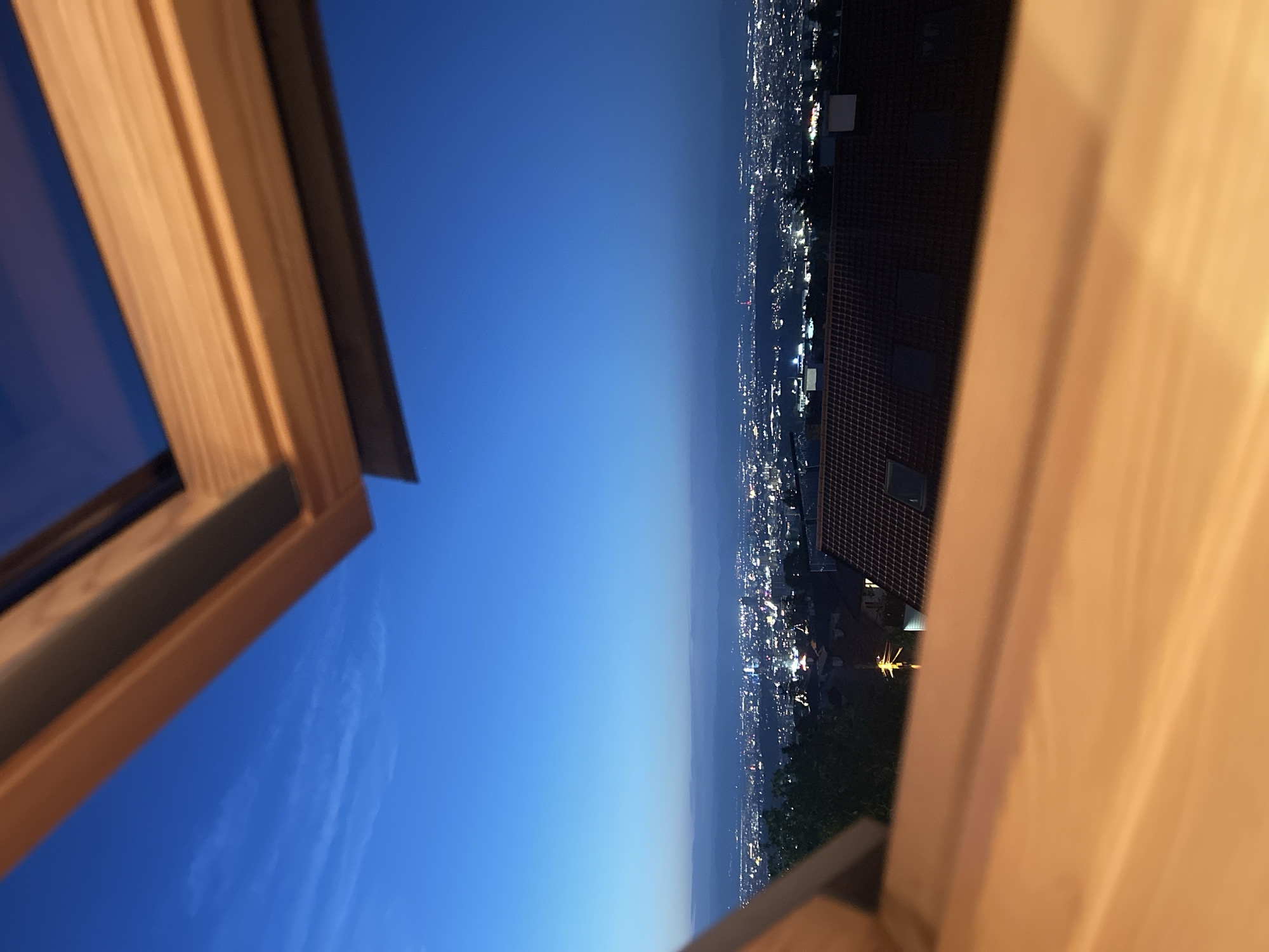 An elevated view of a city skyline at dusk is captured through a wooden-framed open window. The sky is a deep twilight blue with wispy white clouds near the horizon. The city lights twinkle below, suggesting a densely populated area. In the foreground, the blurred wooden frames of the window create a natural frame for the scene. Some rooftops of houses are visible in the mid-ground. The overall impression is one of a serene evening view overlooking a city from a high vantage point.