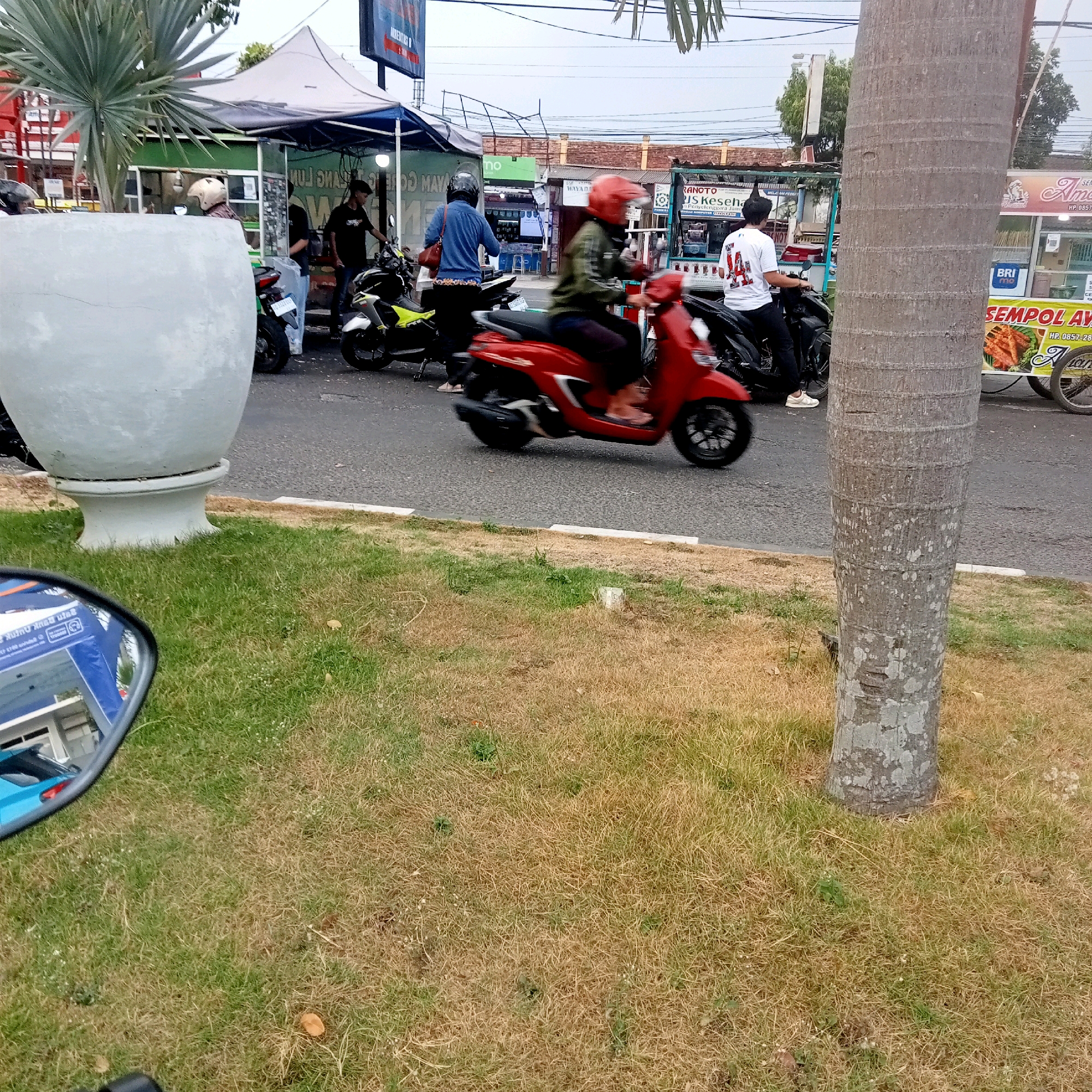 The image captures a street scene in Karanganyar, Indonesia, likely in the vicinity of Alun-Alun Karanganyar. In the foreground, dry grass and a large, light-colored planter are visible. A motorcycle mirror reflects a blue and white building. Beyond the grass, a paved road hosts several people and vehicles. A person on a red scooter with a red helmet is in motion, while another person in a white shirt with the number "14" on it sits on a black motorcycle. Several other motorcycles are parked or being ridden. Tents and food stalls line the side of the road, with signs suggesting food vendors and possibly commercial services. A large palm tree trunk is prominently positioned on the right side of the frame. The background shows buildings and overhead power lines, indicative of an urban environment under an overcast sky. The scene depicts a typical day with people engaged in daily activities on a busy street.