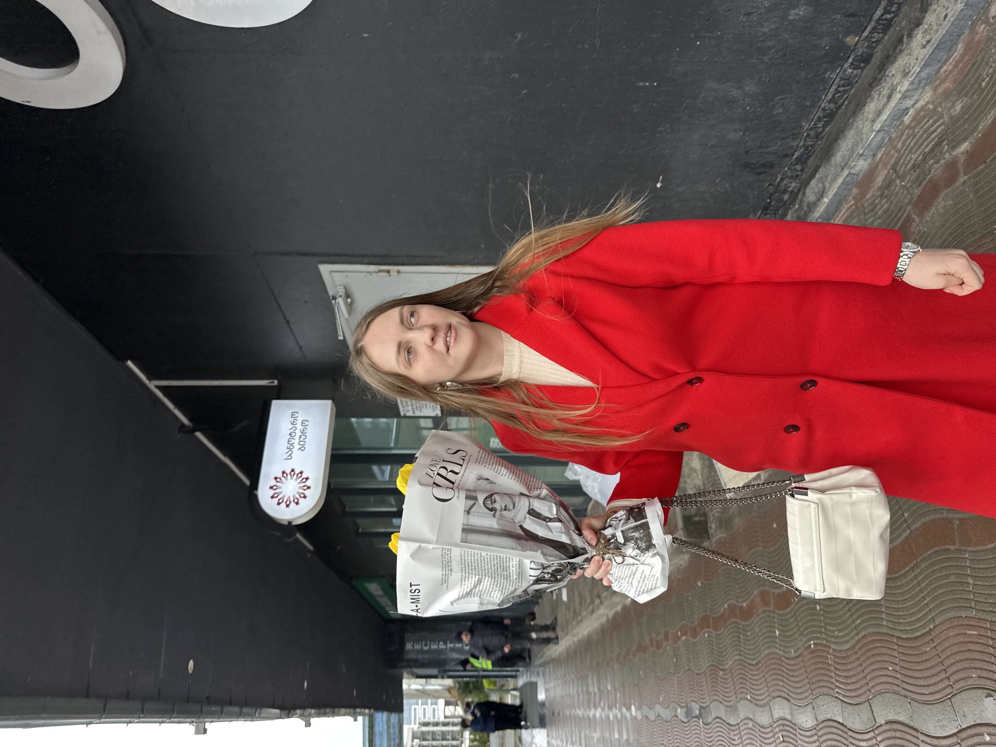 A young woman in a red coat stands on a sidewalk in Batumi, Georgia, holding a bouquet of yellow flowers wrapped in newspaper. She is wearing a watch and has a white purse with a chain strap over her shoulder. The sidewalk has a wavy pattern. Behind her is a black building with a white sign in Georgian script and a white illuminated element. People walk in the background.