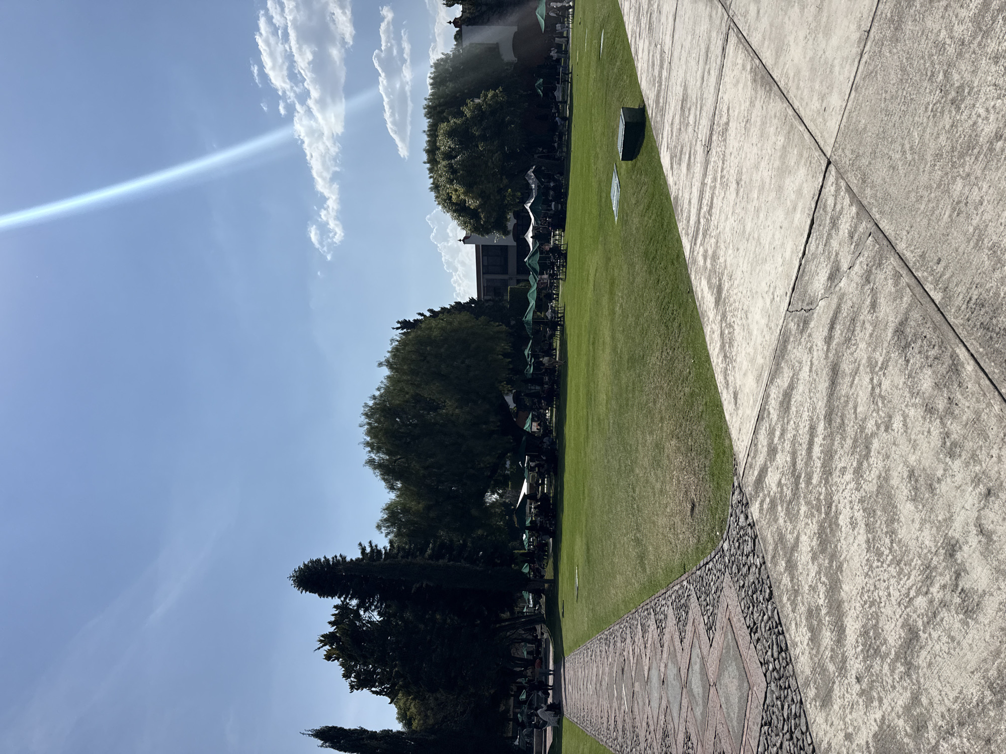 The image shows a sunny outdoor scene in San Andrés Cholula, Mexico. A manicured lawn extends toward a building partially obscured by trees. Numerous green umbrellas suggest an outdoor dining or gathering area. A concrete walkway with textured paving stones borders the lawn. The sky is blue with scattered clouds and a visible lens flare. The perspective is slightly skewed, with the walkway dominating the foreground.