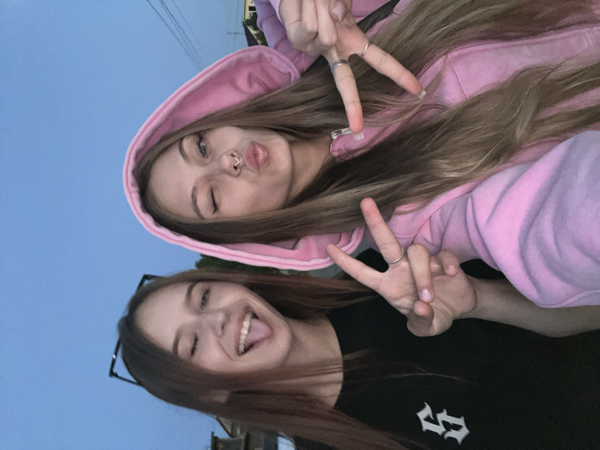 Two young women are posing for a selfie, likely outdoors in Фоки, Russia, given the context. The woman on top is wearing a light pink hooded sweatshirt and has long, light brown hair. She is winking with her left eye and puckering her lips, making a peace sign with her right hand. She has a septum piercing. The woman below is wearing a black shirt with a white design and has long, brown hair. She is smiling broadly with her tongue sticking out and also making a peace sign with her right hand. The sky is a muted blue color, indicating either early morning or late evening. There are also buildings in the background.