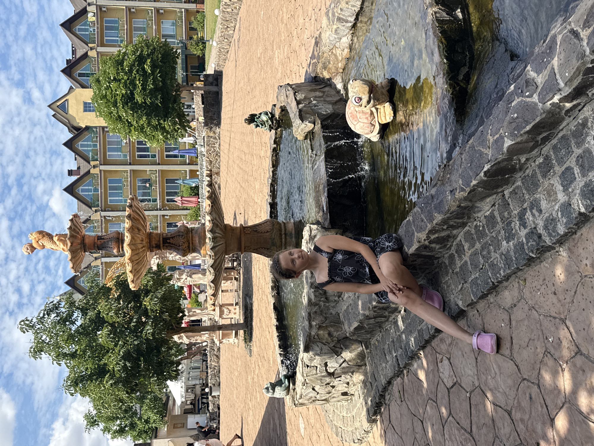 A young girl with brown hair tied back sits on the edge of a stone fountain. She is wearing a dark sleeveless dress with a floral pattern and pink clogs. Her legs are crossed at the ankles, and her arms are resting on her lap. The fountain is made of rough-hewn stones and features a statue of a turtle in the water. In the background, a multi-story yellow building with arched windows and balconies is visible. Several trees and other decorative statues are also present in the courtyard. The ground is paved with decorative stone tiles. The sky is cloudy. The overall setting appears to be an outdoor courtyard or recreational area.