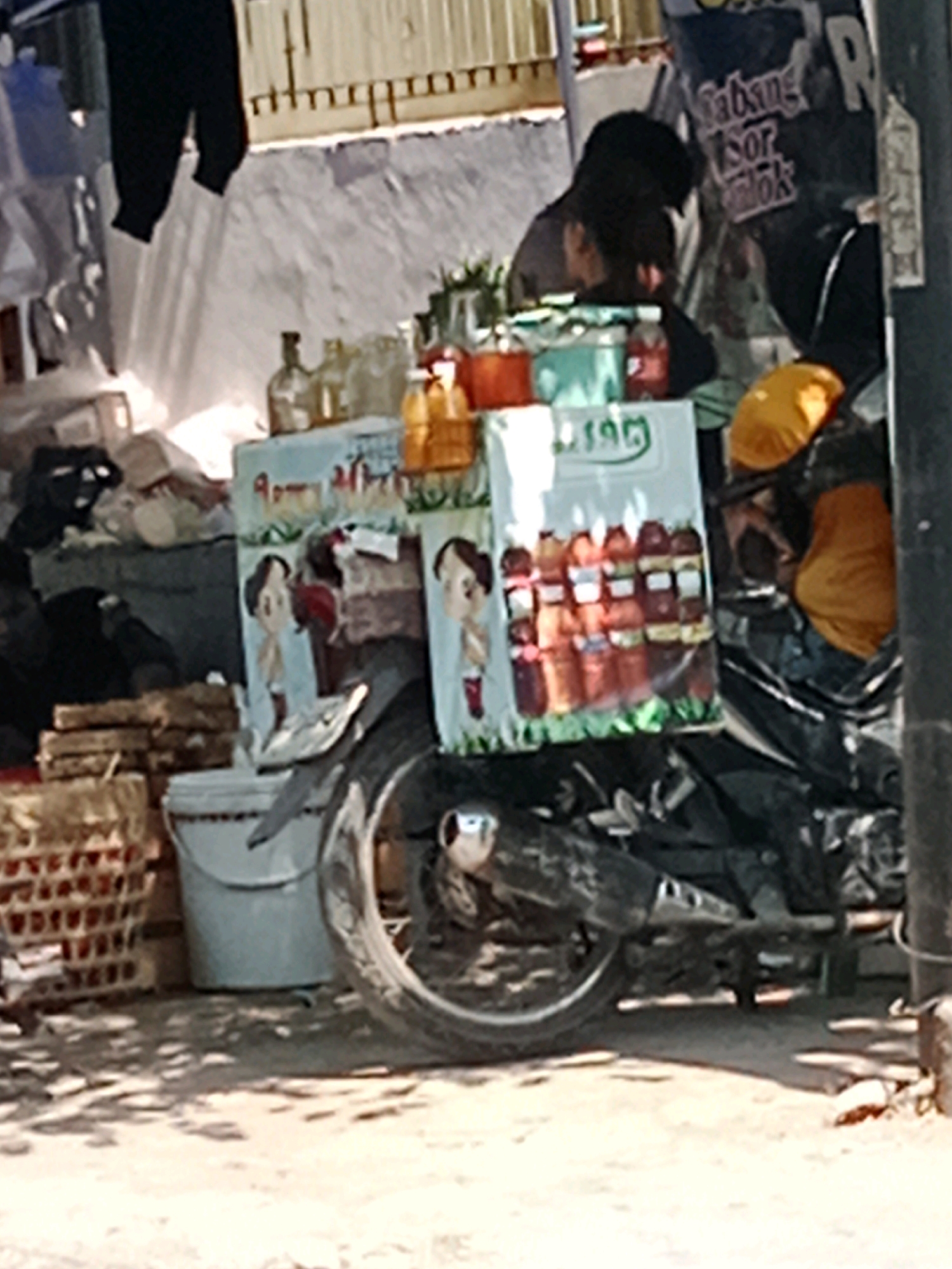 The image depicts an outdoor scene in Banaran, Indonesia, likely a street vendor's stall. A motorcycle is parked in the foreground, partially obscured by various items. Behind the motorcycle, a display of bottles, possibly beverages, is arranged. Two cartoon-like figures are visible on a board above the bottles. In the background, a person is partially visible, seated on or near the motorcycle. The environment appears somewhat cluttered, with crates, a bucket, and hanging items suggesting a temporary setup. The lighting suggests it is daytime.