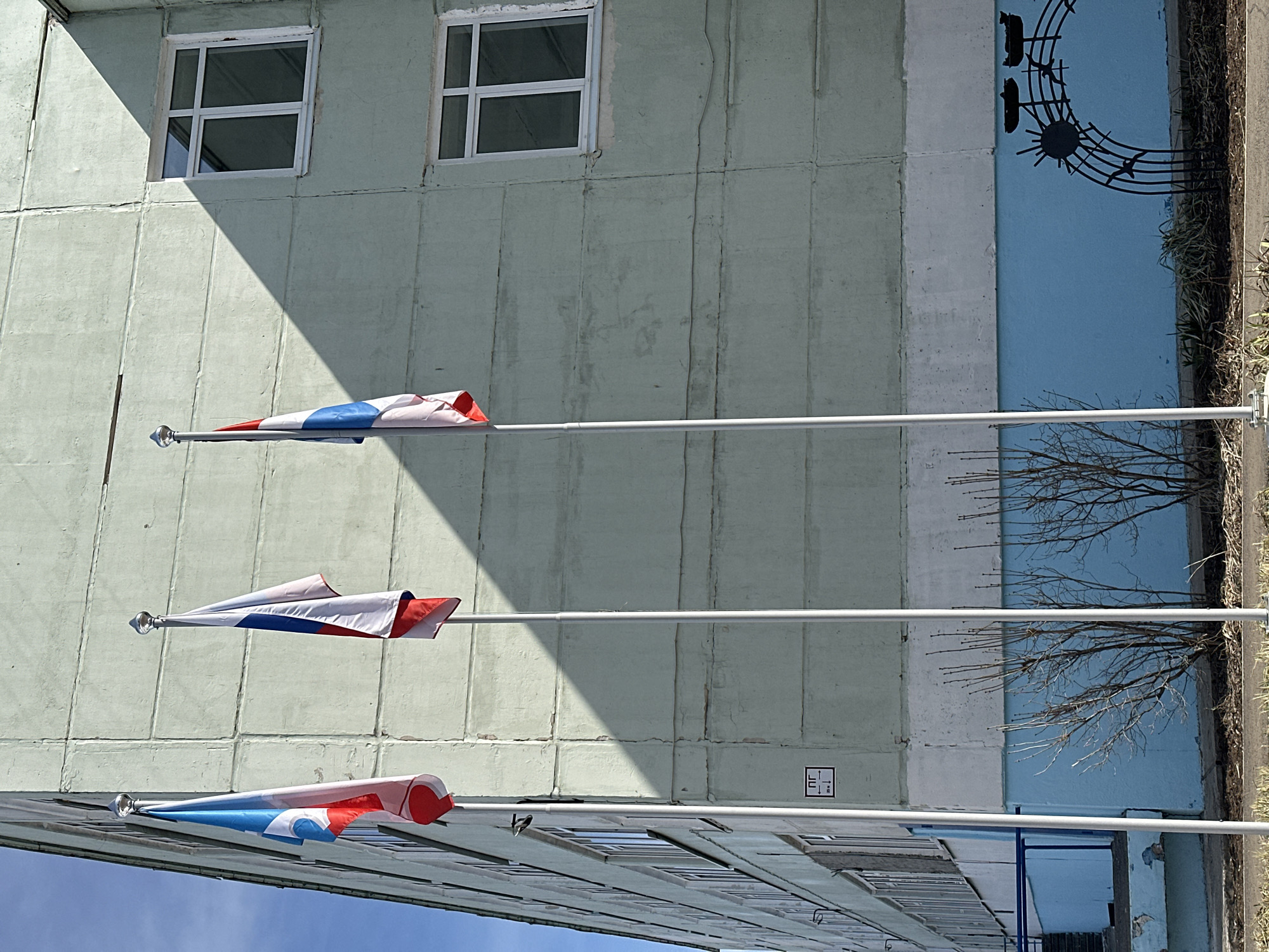 The image shows a building in Chaikovsky, Russia. The building is light green and has two windows. Three Russian flags are mounted on poles attached to the building. The flags are red, white, and blue. A blue mural with a musical note design is on the right side of the building. Bare trees and a sidewalk are visible on the right edge of the image.
