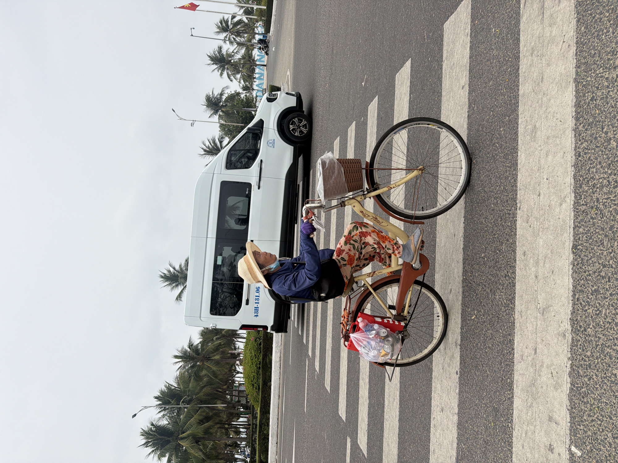 A person is seen riding a classic-style bicycle across a white-striped crosswalk on an asphalt road in Đà Nẵng, Vietnam. The individual, who appears to be elderly, wears a straw hat, a blue jacket, floral-patterned pants, and a light blue face mask. The bicycle features a cream-colored frame, a woven basket on the handlebars containing a white plastic bag, and a transparent plastic bag filled with aluminum cans and plastic bottles secured to the rear rack, suggesting the rider may be collecting recyclables.
In the background, a white van with the license plate "43H-131.06" is visible. A line of tall palm trees stretches along the side of the road. A sign with the text "DANANG" is partially visible among the trees, confirming the location. In the distance, a red and yellow flag, characteristic of the Vietnamese national flag, flies from a pole. The scene is illuminated by bright, diffused daylight, indicating an overcast sky.