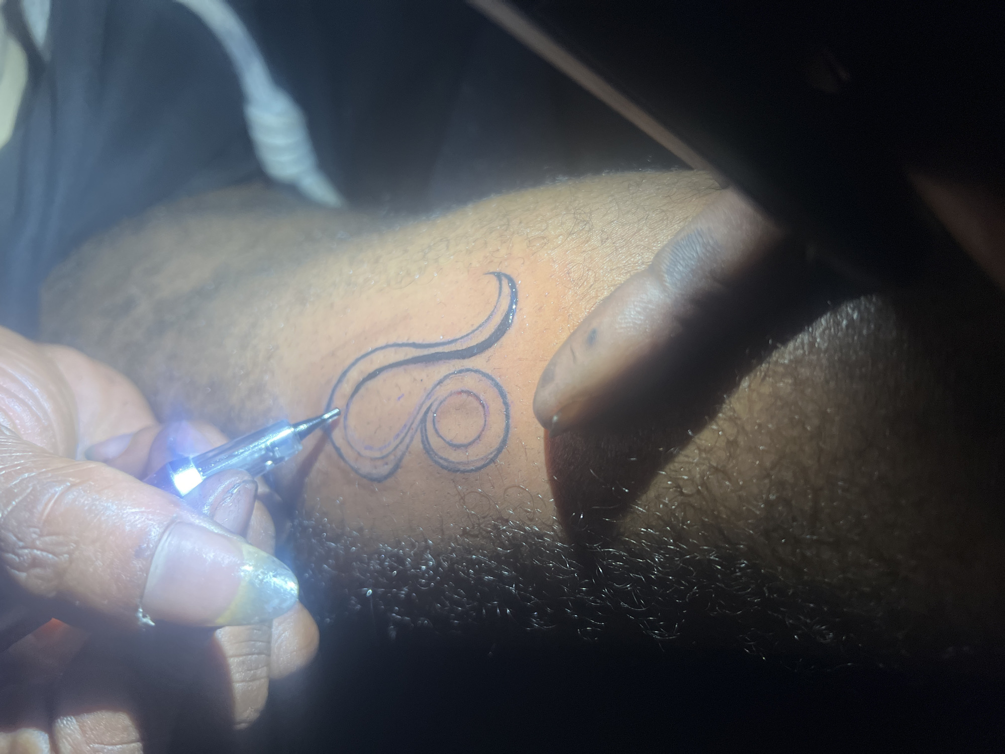 The image shows a close-up view of a tattoo being applied to skin. A tattoo needle, attached to a device with a light, is in contact with the skin, creating the outline of a Leo zodiac symbol. The skin appears to have dark hair follicles. Another hand is partially visible, holding the skin taut. The lighting suggests the image was taken in a dimly lit environment, possibly a tattoo parlor or a personal setting. The location in Gura Top, Nigeria, cannot be determined from the visual information provided in the image.