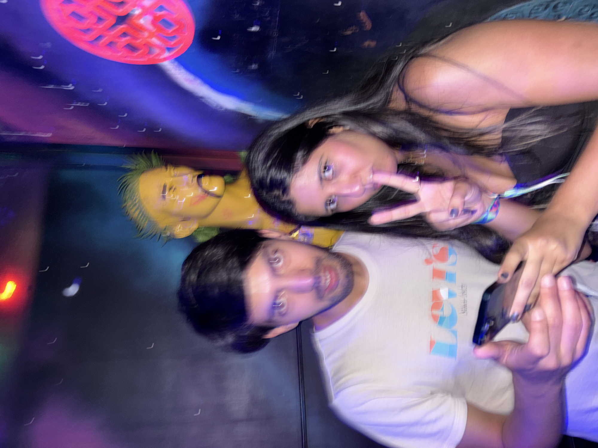 A blurry photograph shows two people in an indoor venue. A man with dark hair and a beard wears a white Levi's T-shirt and looks at the camera while holding a smartphone. A woman with long, dark hair is next to him, holding up a peace sign. Behind them is a yellow-skinned mannequin head with green hair and a wall with colorful decorations, including a red, geometric object, and what appears to be artwork of a planet or celestial body. Small, blurry white spots are scattered throughout the background. The image is blurry and tilted. The location is possibly in São Paulo, Brazil, though this is not definitively confirmed by visual elements.