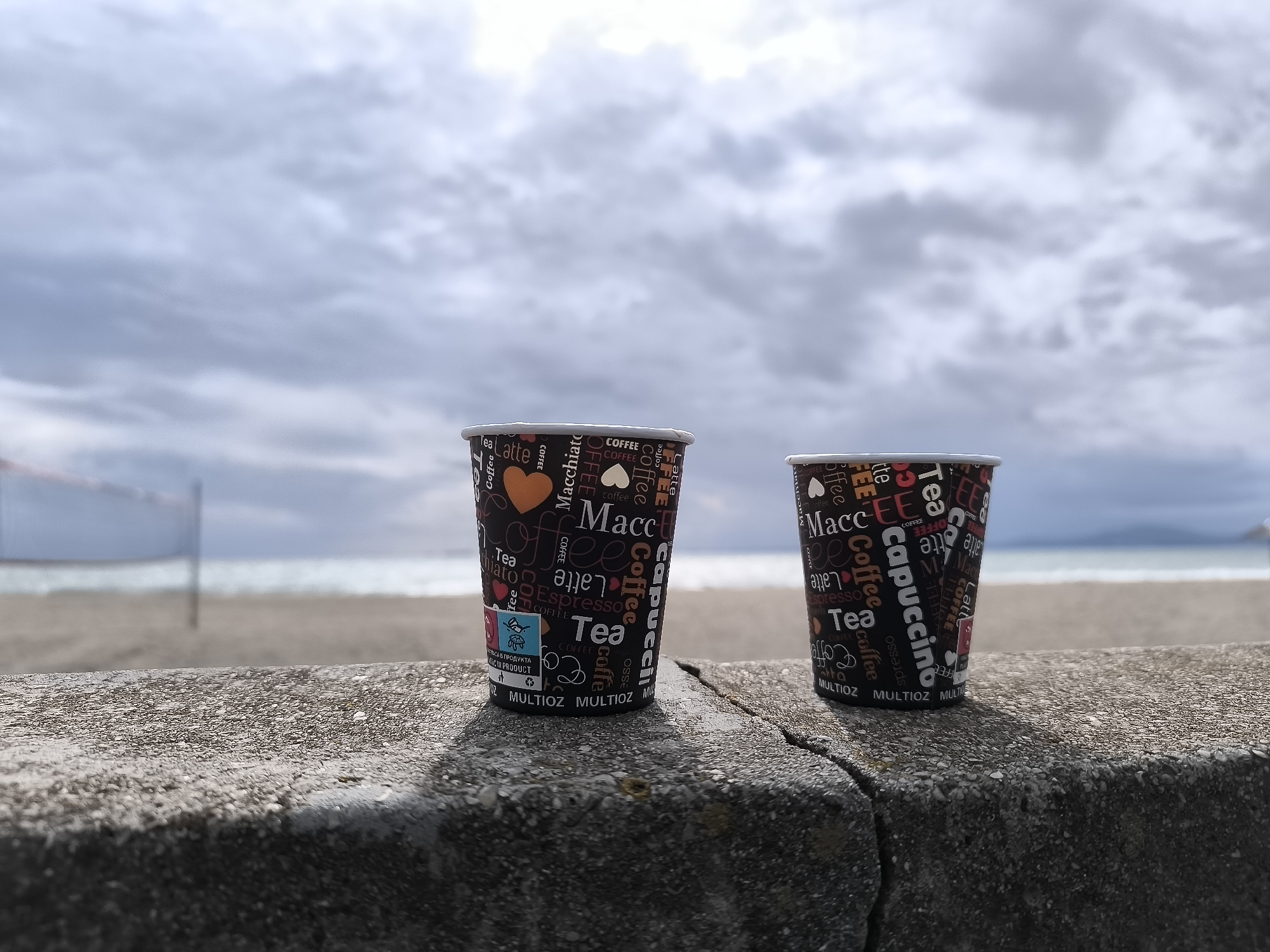 The photograph shows two paper cups of coffee sitting on a concrete barrier. The cups have a black background with white and red text and graphics related to coffee and tea, including words like "Macchiato", "Latte", "Espresso", and "Cappuccino". An orange heart is visible on the left cup. The barrier is gray and has a crack running across it.
In the background, there is a sandy beach, the sea, and a cloudy sky. A volleyball net is faintly visible on the beach. Based on the location and the presence of the sea and beach, this image was taken in Burgas, Bulgaria.