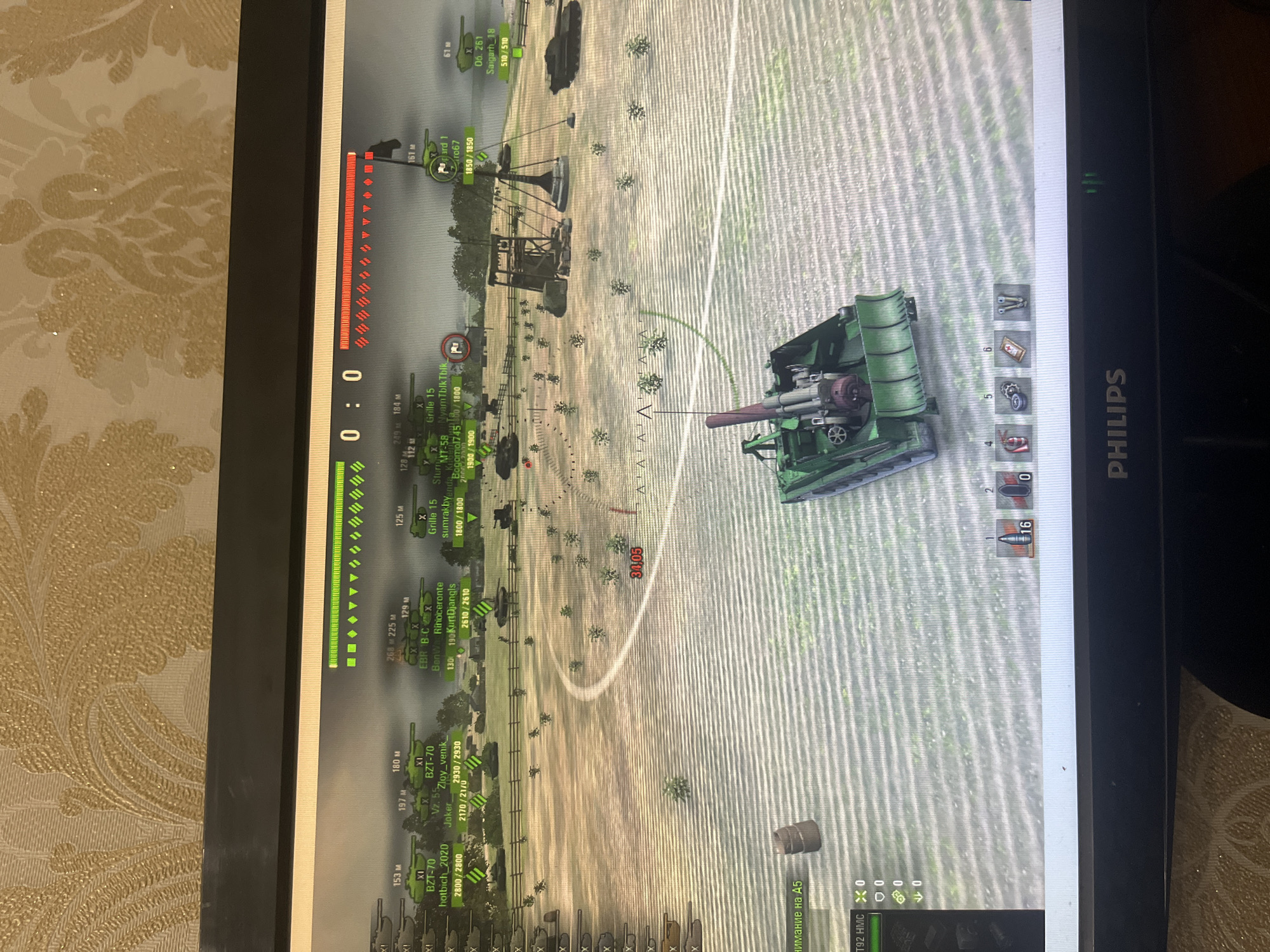The image shows a screen displaying a game, likely a tank combat simulation, with a green artillery vehicle in the foreground aiming towards a distant structure. The environment appears to be a field with scattered vegetation. The in-game user interface displays player information, health bars, timers, and a mini-map. The timestamp reads 0:0. There's no indication of a location in Kaspiysk, Russia, or any people present in the scene.