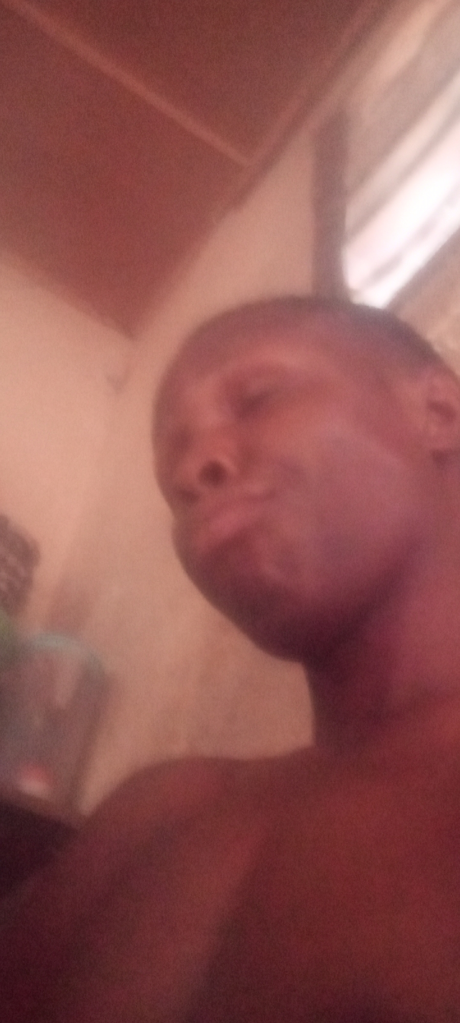 The image is a blurry, close-up portrait of a person, likely male, with dark skin, captured from a low, upward angle. The individual's head is tilted slightly, and their gaze appears to be cast downwards or their eyes are closed, with a subtle pout or relaxed expression on their lips. They appear to be shirtless, with a bare shoulder visible in the lower right.
The overall lighting is dim and warm, suffusing the entire frame with reddish-brown and soft pink tones. This low-light environment suggests an indoor setting, possibly during the evening or night. The background is extremely blurred and indistinct, showing only vague shapes and variations in the warm color palette, which could be walls or other interior architectural elements. No specific objects or identifying features of a room are visible, nor is any text.
The photograph conveys a sense of intimacy and introspection due to its close cropping, soft focus, and subdued lighting. The lack of sharp detail and the warm hues create an ethereal, slightly pensive mood. The image is likely a self-portrait or a casual snapshot taken in Sabon Tasha, Nigeria.