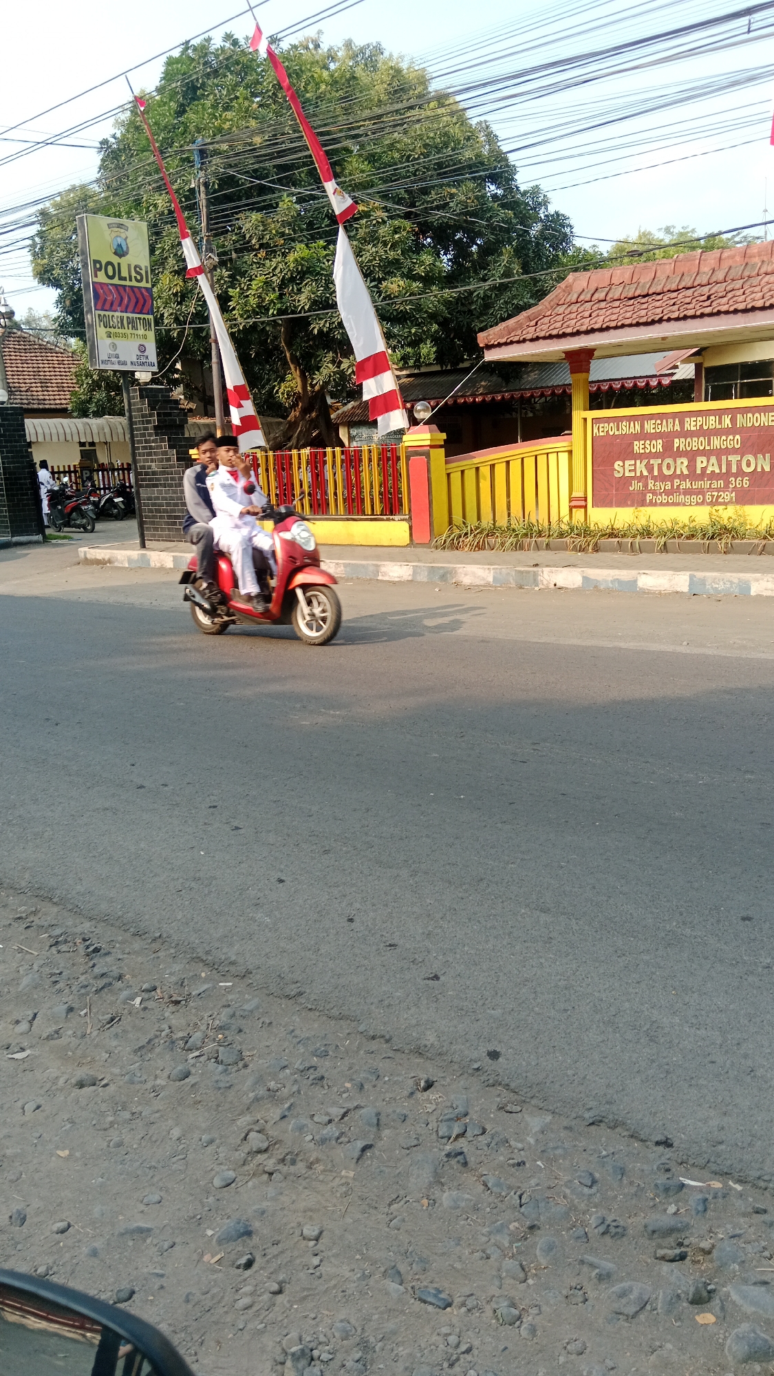In the city of Patunjungan, Indonesia, a red scooter with two occupants is seen driving on an asphalt road. The rider is a male, and he is wearing a white uniform with a cap. The passenger is a male, wearing a dark blue jacket and dark pants. The scooter is in motion, suggesting the subjects are traveling.
Behind the scooter, there is a police station with a sign that reads "POLISI SEKTOR PAITON". The police station has a yellow fence with red accents. Indonesian flags, red and white, are displayed on poles in front of the station. Trees and power lines are visible in the background. The sky is clear and bright, indicating daylight. The road appears to be in fair condition, with some gravel and debris on the side. The overall environment suggests a street scene in a relatively populated area, likely during the day.