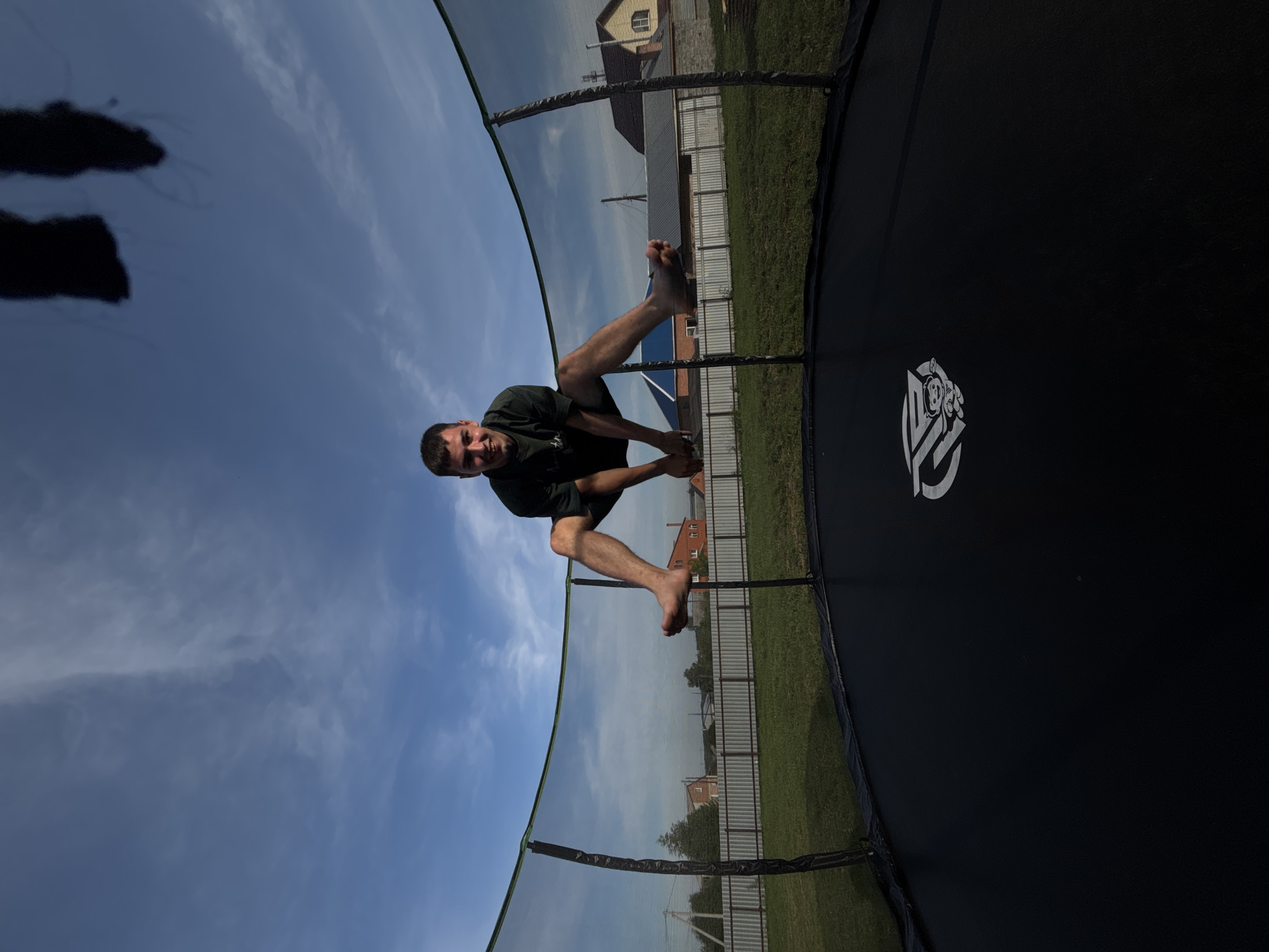 A young man is captured mid-air, jumping on a trampoline on a sunny day in Vishnevka, Russia. He is wearing a dark green t-shirt and dark shorts, with his legs spread wide as he propels himself upwards. His bare feet are visible, one slightly ahead of the other. He is smiling at the camera with a happy expression.
The trampoline has a black jumping surface with a white logo in the center, featuring a cartoon character. The trampoline is surrounded by a green safety net, held up by dark poles. The background shows a typical rural landscape, with a well-maintained green lawn, a white picket fence, and several houses with tiled roofs, suggesting a residential area. The sky above is a clear blue with some scattered white clouds, indicating good weather conditions. The overall scene depicts a moment of outdoor recreation and enjoyment.