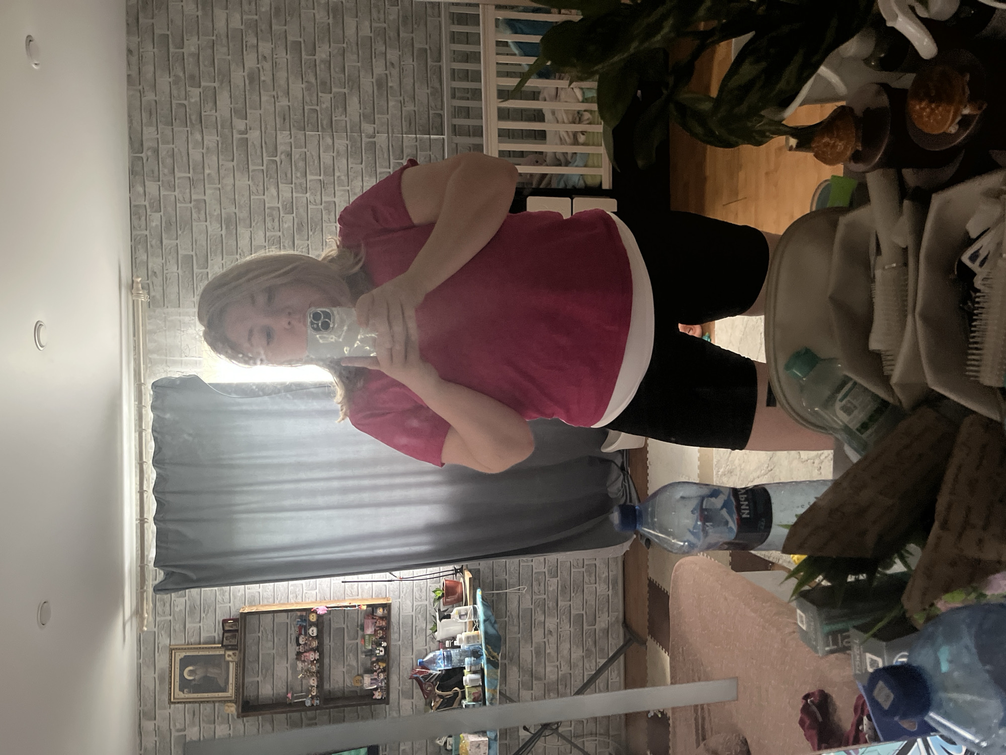 A woman is taking a selfie in a mirror. The location, though a mirror image, appears to be an interior space with a brick-patterned wall. Other visible items include a window with a grey curtain, framed pictures on the wall, a small table with various objects, a water bottle, and plants. The woman is wearing a pink shirt and black shorts.