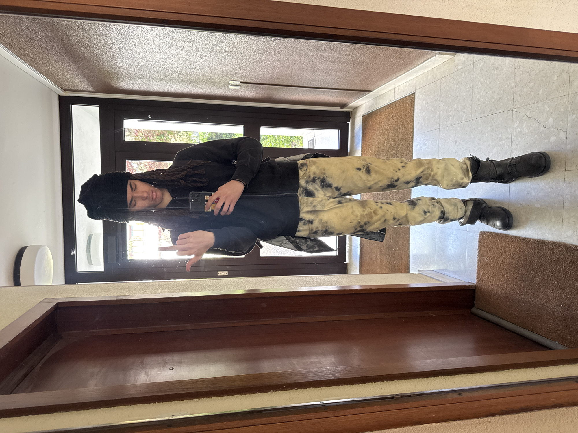 A person wearing a black beanie, a black hoodie, and patterned pants is standing inside a doorway in Les Ulis, France, taking a photo of themselves in a mirror. The doorway leads to an exterior scene visible through a glass door. The floor is tiled, and there is a brown mat near the door. The door frame is brown wood. The person is holding a phone and making a hand gesture.