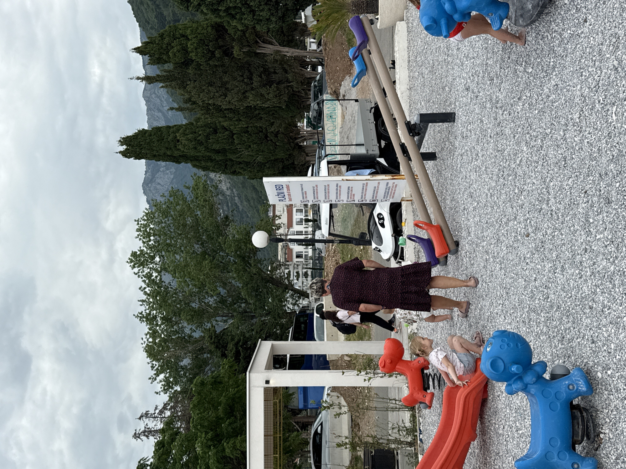 A playground area in Budva, Montenegro, is populated by children and adults. A young girl sits on a red plastic slide, next to a blue plastic bouncy horse. Another child is visible near a blue bouncy horse in the background. An adult woman in a dark-colored dress walks past the playground equipment with a young girl.
Behind the playground, a modern white pergola structure stands, and cars are parked. A sign labeled "PLAZNI RED" in white lettering is visible in the middle ground. The background includes trees, buildings, mountains, and a cloudy sky.