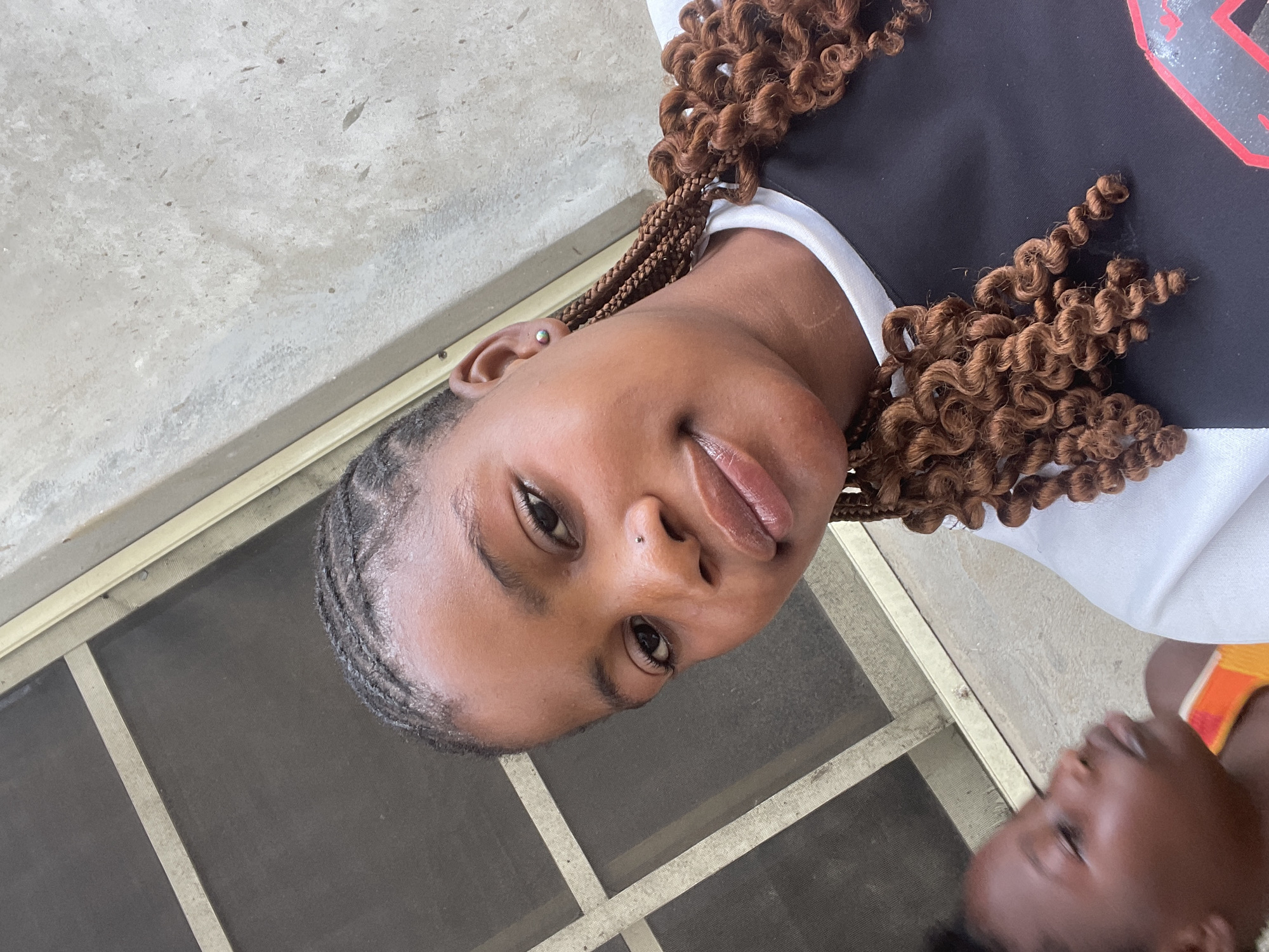 The image presents a close-up portrait of a young person, likely a teenager or young adult, in an outdoor or semi-outdoor setting in Badagry, Nigeria. The main subject has brown skin, dark eyes, and a relaxed, possibly slightly smiling expression, looking directly at the camera. Their hair is styled with cornrows at the front and sides, transitioning into voluminous, curly brown braids or extensions. They have a small stud piercing on their left nostril and another on the helix of their left ear.
The individual wears a black top with white trim, which features a partial red graphic design, possibly a letter or logo, visible on the upper left side of the black fabric. In the background, a light-colored concrete or tiled surface is visible, along with a screen door or window frame with a metallic finish. In the lower right corner, another person's head is partially visible, also with brown skin, seemingly looking upwards and wearing a garment with orange and yellow accents. The natural lighting suggests the photograph was taken during the daytime. The scene appears casual and captures an everyday moment.