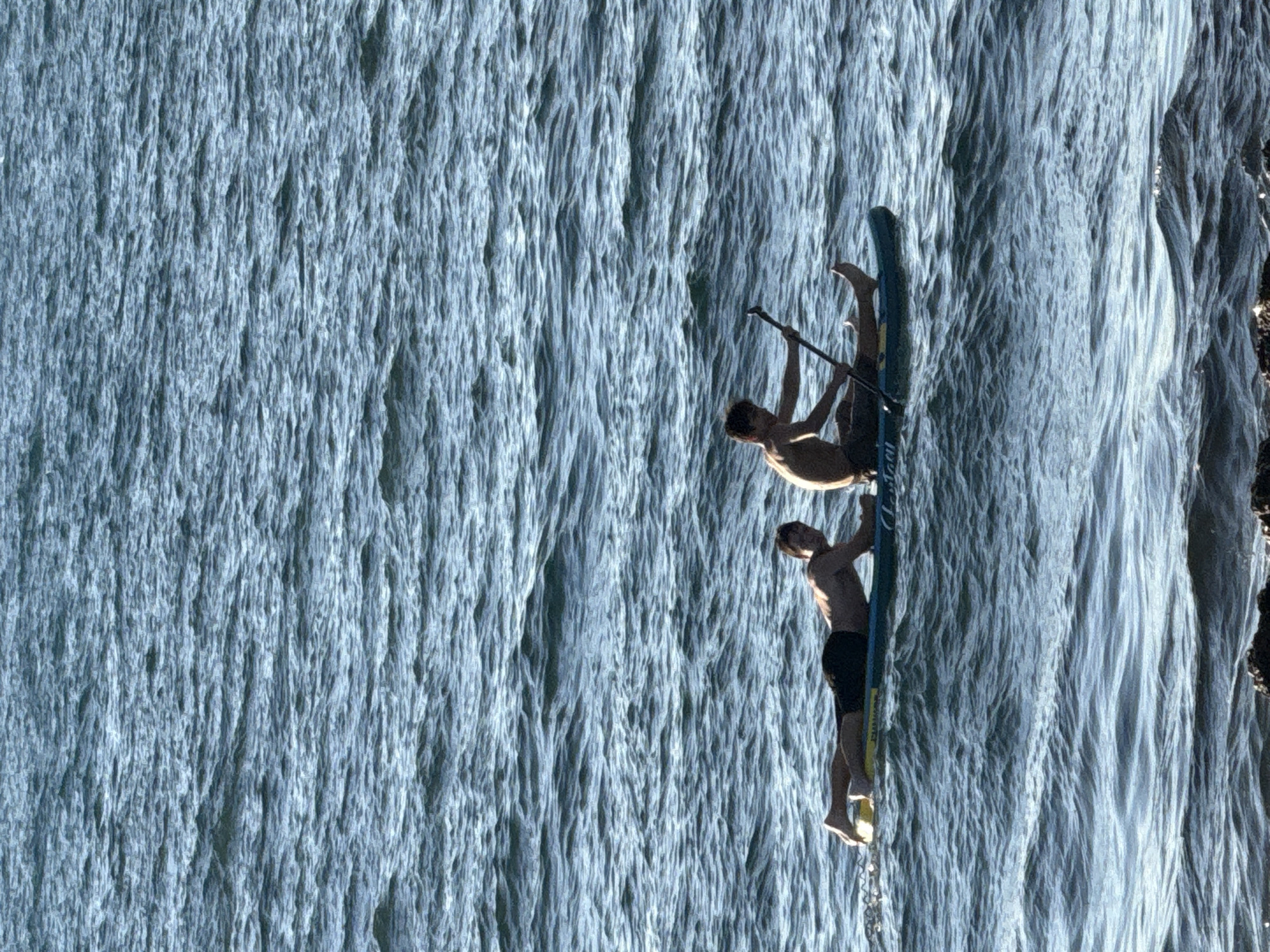 Two young men are paddleboarding on a body of water. The man in the foreground is sitting on the paddleboard, holding a paddle. The man in the background is lying on his stomach on the paddleboard. The water is blue and choppy, with small waves. The sky is not visible. The scene appears to be a recreational activity in an outdoor setting.