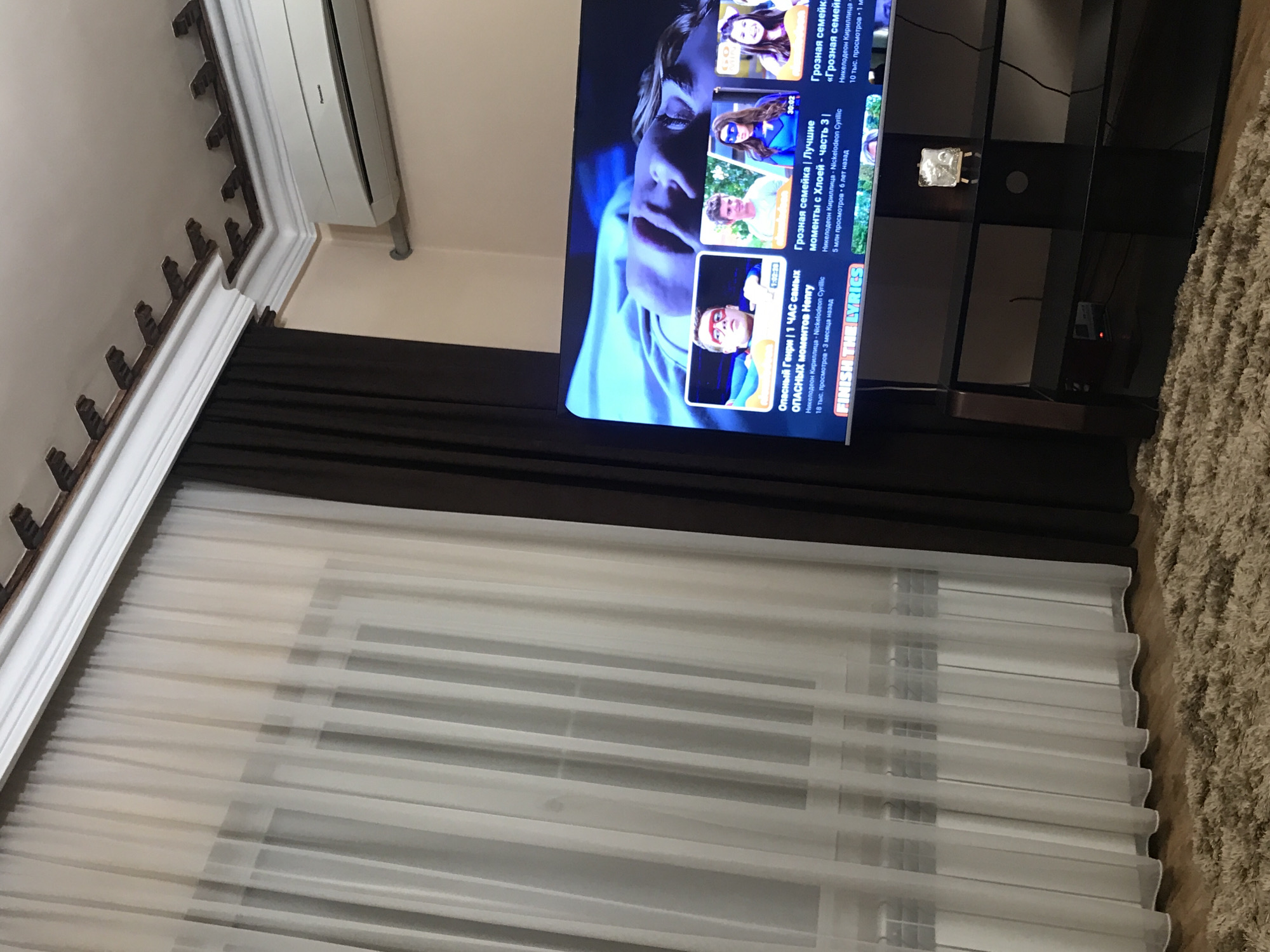 The image shows the interior of a room, likely in a home, with a television on a stand, along with curtains covering a window. The television is displaying content from YouTube. The curtains are a combination of dark brown and white sheer fabric. A beige carpet is visible on the floor. The wall has a white molding and an air conditioning unit is mounted at the top. Without any identifiable location-specific features within the image, determining its precise location in Yerevan, Armenia is not possible.