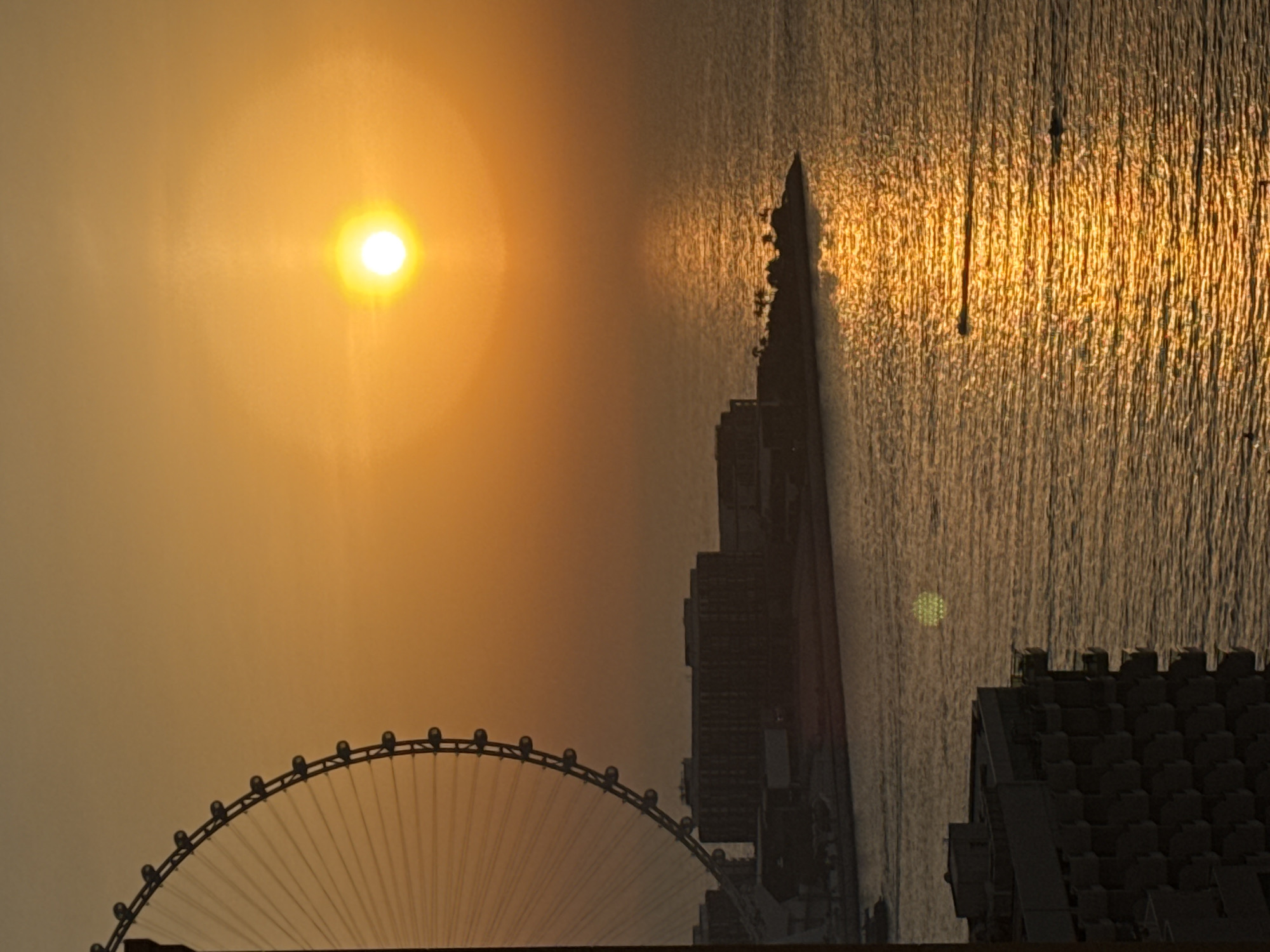 The image captures a sunset over the Arabian Gulf in Dubai, United Arab Emirates. The foreground features the side of a multi-story building, with balconies visible. In the mid-ground, a large Ferris wheel, likely the Ain Dubai, stands prominently on the left side, its structure silhouetted against the warm, orange sky. A long, low-rise building complex is situated on land extending into the water. The water itself reflects the bright sun, creating a shimmering golden path. Two small boats are visible on the water, one leaving a wake. The overall scene is serene, dominated by the warm tones of the sunset and the expansive seascape.