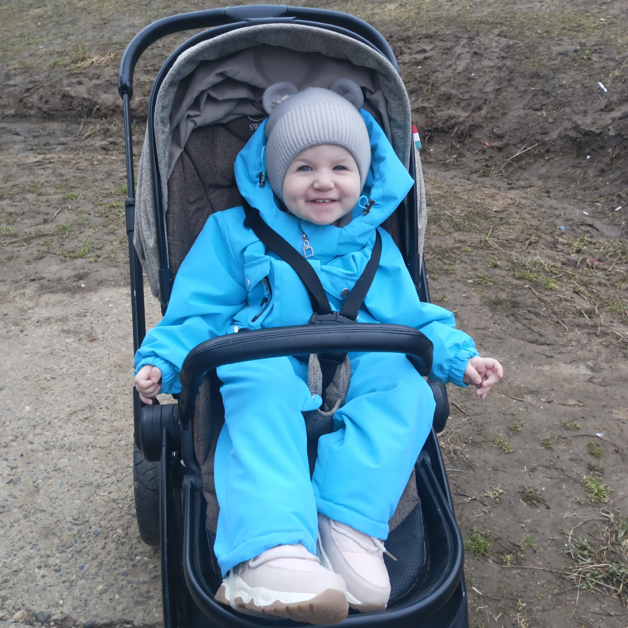 A young child is seated in a black stroller, smiling, against a backdrop of a muddy, unpaved area with some sparse vegetation. The child is wearing a bright blue snowsuit, a gray knit hat with bear ears, and light pink shoes. The stroller has a neutral-colored interior lining and a black safety bar in front of the child. The presence of a small flag with green, white and red stripes suggests the location is within the city of Chaikovsky, Russia.