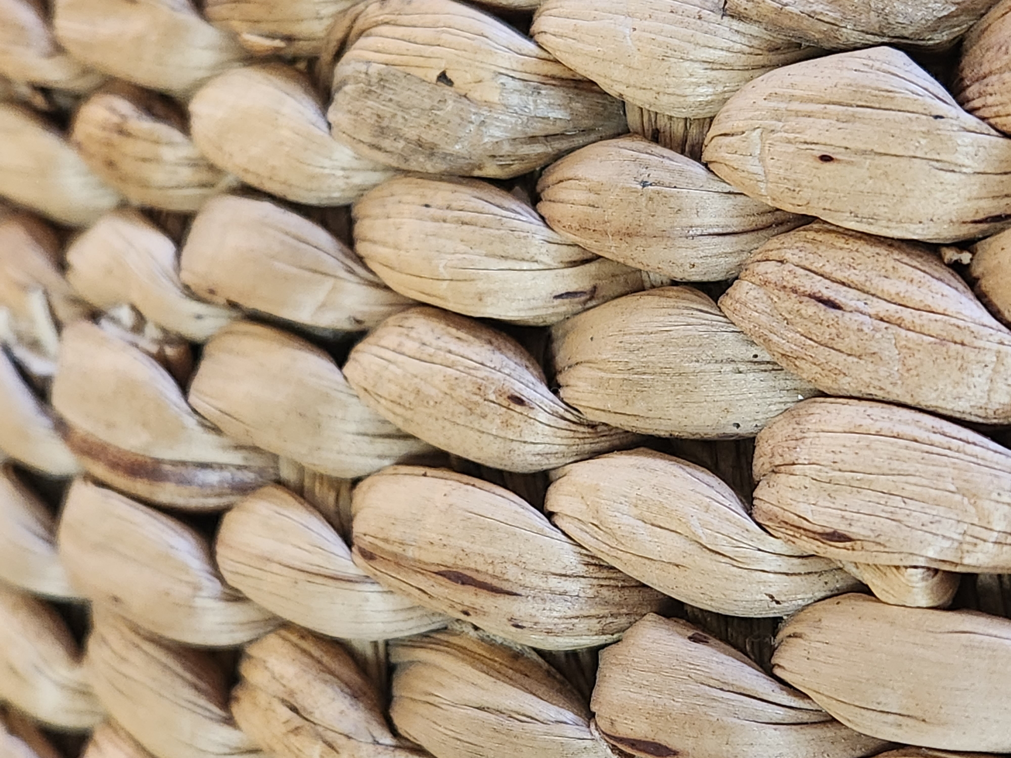 This is a close-up, macro photograph of a woven basket. The subject is the texture and pattern of the woven material, which appears to be made of dried, natural fibers like straw or rattan. The fibers are tightly braided and twisted, forming a diagonal pattern across the frame. The color palette is dominated by natural, earthy tones of beige, light brown, and cream, with some darker brown or reddish-brown streaks and marks on the fibers, suggesting age or natural variations in the material. The lighting is soft, highlighting the textures and the three-dimensional form of the weave. The shallow depth of field blurs the background and foreground, focusing attention on the intricate details of the basket's construction. There is no visible text, people, or discernible activity taking place; the image solely focuses on the material and texture of the object. The setting is likely domestic or artisanal, given the object is a basket. The image conveys a sense of natural materials and craftsmanship.