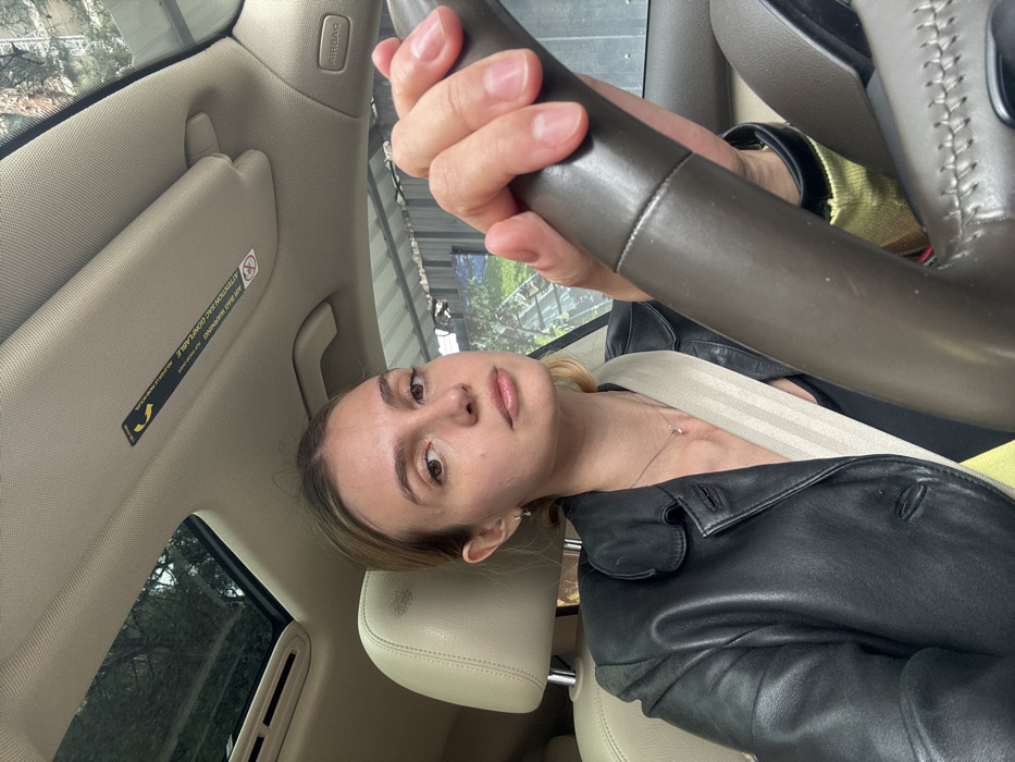 The image depicts a female individual positioned in the driver's seat of a vehicle, capturing a selfie. The subject, with light brown hair pulled back, possesses light skin and maintains a neutral gaze towards the camera. She wears a black leather jacket over a dark garment, secured by a cream-colored seatbelt diagonally across her chest. A delicate silver necklace with a pendant and small silver stud earrings are present. Her left hand is clasped around the dark brown, possibly leather-wrapped, steering wheel. The car's interior features beige/tan upholstery, a visible sunroof reflecting tree canopies, and an "AIR BAG" warning label affixed to the overhead visor. The exterior environment, visible through the vehicle's windows, comprises trees, a structure with a corrugated metal roof, and a large visual display featuring a green landscape. This urban setting, characterized by green spaces and architectural elements, is consistent with typical environments found within Tbilisi, Georgia.
