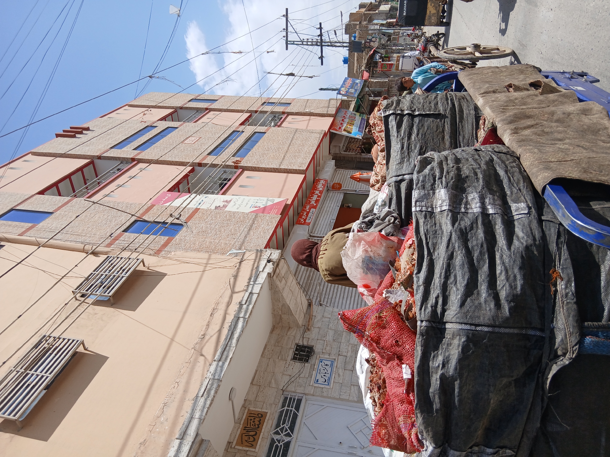 The image depicts a street scene in Quetta City Tehsil, Pakistan, with a building on the left featuring multiple floors and windows with blue glass. A variety of objects are stacked on a cart in the foreground, covered by a tarp and red netting. Overhead, power lines crisscross the sky. Further into the scene, storefronts are visible.