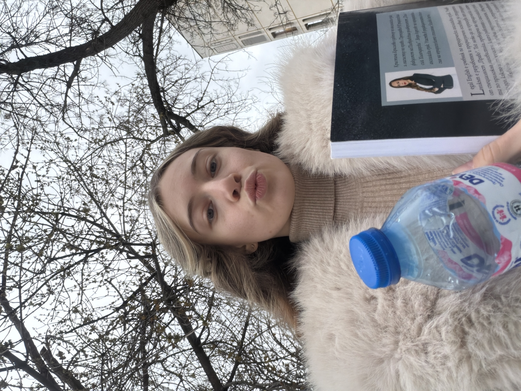 A young woman poses for a selfie outdoors. She is wearing a beige turtleneck sweater and a fluffy, off-white coat. She is holding a book and a blue water bottle. The book has a photograph of a woman on the cover, along with Russian text. Behind her, bare tree branches fill the frame, and a building is partially visible in the background. The image does not indicate the location is in Plovdiv, Bulgaria.
