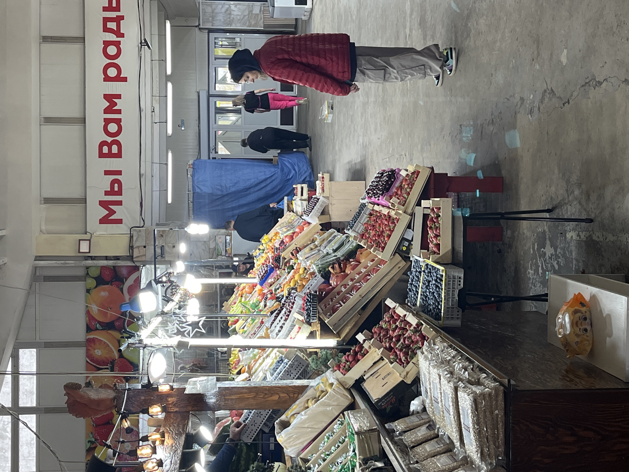 The image shows the interior of an indoor market. A person in a red jacket and grey pants is standing near a fruit and vegetable stand. The stand is stocked with various fruits and vegetables in wooden crates. Other people are visible in the background near the entrance of the market. A sign with red Cyrillic text hangs from the ceiling on the left. There are several lights and decorations hanging from the ceiling, with an image of fruit behind. The market appears to be busy. The city and region are unknown.