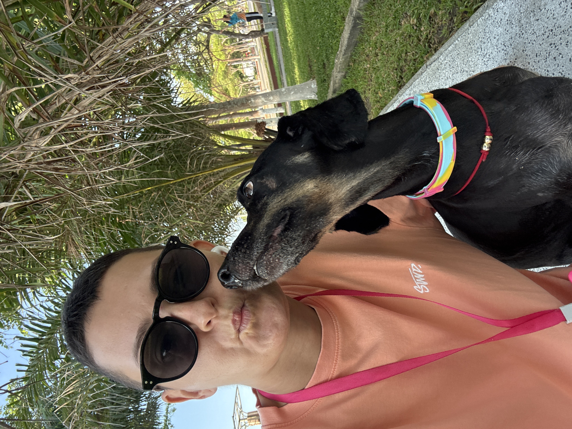 In Nha Trang Ward, Vietnam, a person with short hair and sunglasses is taking a selfie with their dog. The person wears an orange t-shirt with a pink lanyard. The dog, a black dachshund, is wearing a colorful collar and a red string with two gold beads. They are sitting on a bench in a park-like setting with palm trees and green grass. In the background, two people are visible, one stretching and the other standing near a railing.
