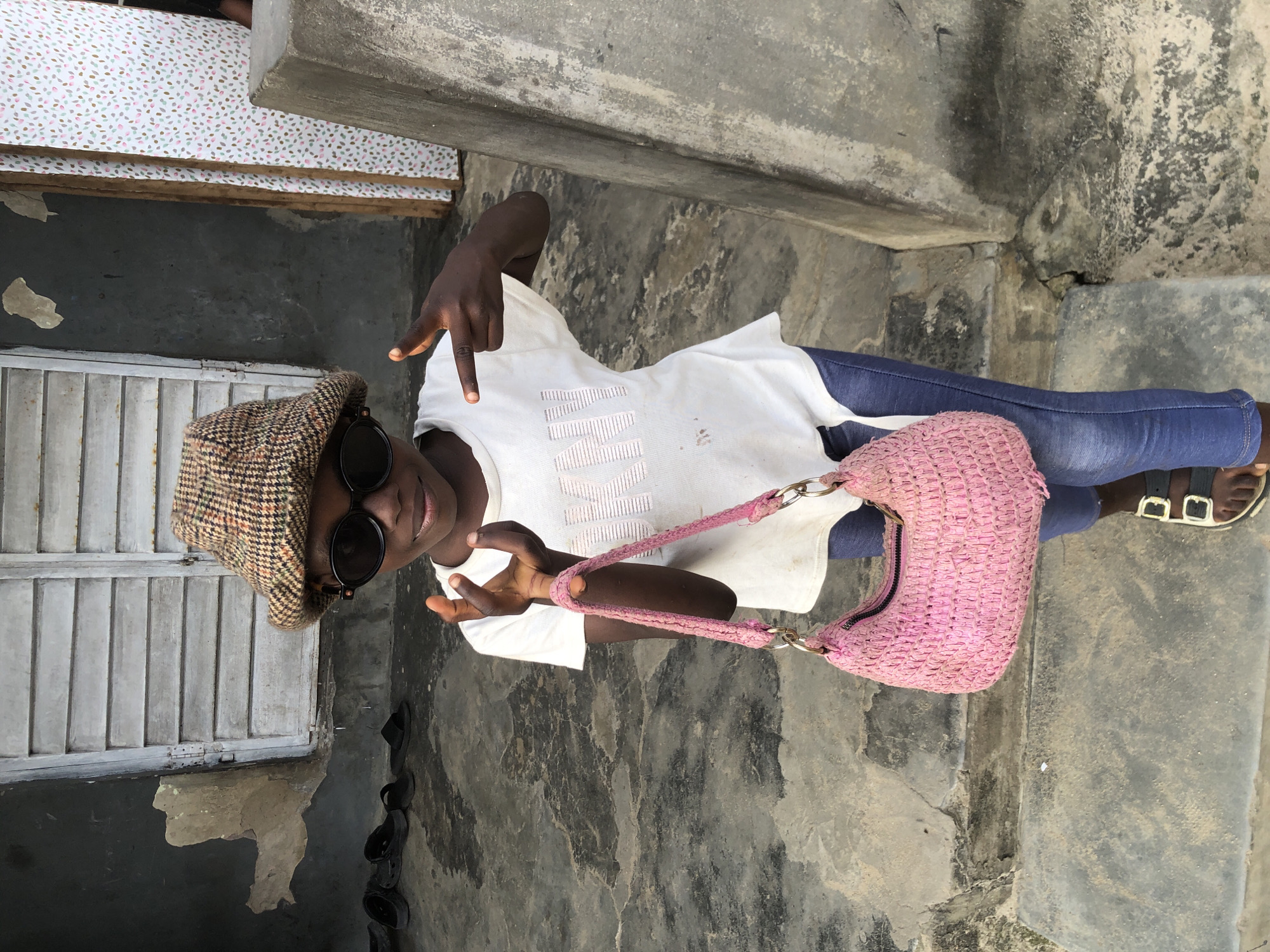 A young Black person wearing a plaid bucket hat and large black sunglasses stands on concrete steps in front of a building in Ado, Nigeria. The person is wearing a white t-shirt with pink text and blue jeans. They are holding a pink woven purse with a chain strap. The person is gesturing with both hands. The background shows a weathered building with gray, slatted doors and a wall with a patterned fabric covering. The ground and steps are concrete, showing signs of wear and dirt. Several pairs of black sandals are visible on the ground to the left. The lighting suggests it is daytime, with shadows cast on the ground.
