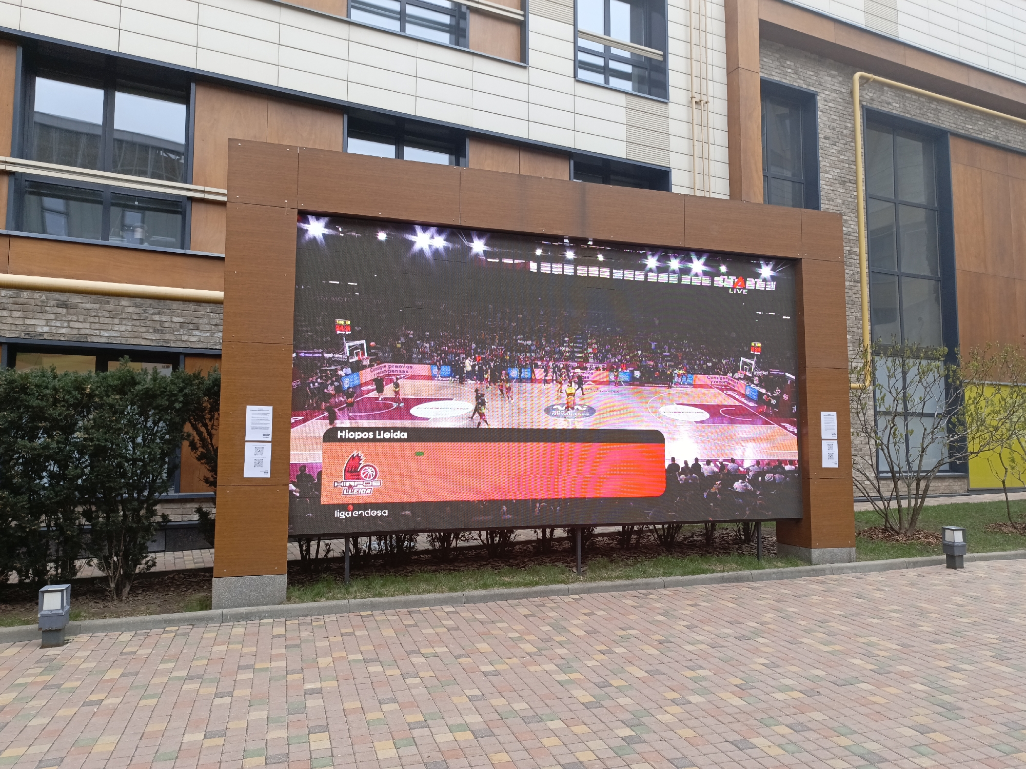 The image shows a large outdoor LED screen displaying a live basketball game. The screen is framed with brown wooden material and positioned on a paved area with brick patterns, likely a public square or courtyard. The broadcast features the "Hiopos Lleida" team and mentions "Liga Endesa". The screen stands in front of a multi-story building with windows and architectural details of cream or beige and brown tones. There are bushes and small trees on both sides of the screen. The sky is not visible, but the lighting suggests it's daytime. The location is Moscow, Russia.