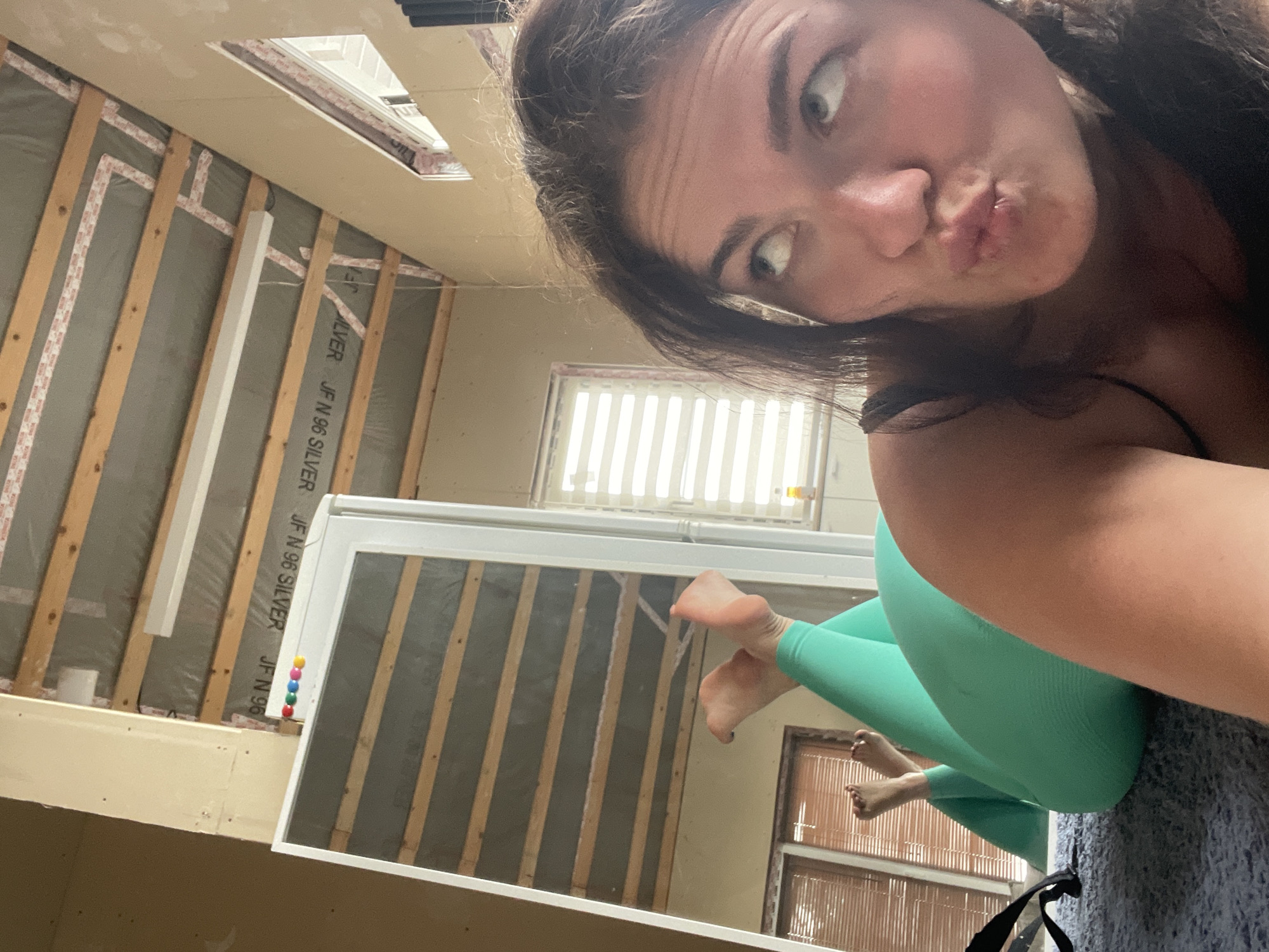 A woman in bright green leggings is taking a selfie in a room under renovation. The walls are partially finished with insulation and wooden beams, bearing the text "JF N 96 SILVER." A skylight is visible in the ceiling. Reflected in a mirror are window blinds. No other information is available to determine the location in the city of Мосметаллоконструкция Онт, Russia.