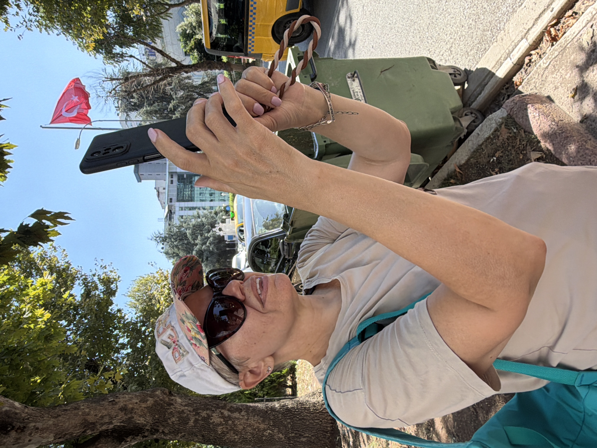 A woman wearing a cap and sunglasses is taking a picture with her phone. She is standing outside, with trees and buildings in the background. A Turkish flag is visible flying from a flagpole. There is a trash can and a yellow vehicle in the scene. The woman is holding a phone in her right hand and her left hand is blurred in motion. She is wearing a light-colored t-shirt and a turquoise bag is slung over her left shoulder. Her fingernails are painted pink. She is wearing a bracelet on her left wrist. The overall impression is that of a candid outdoor photograph being taken on a sunny day. The presence of the Turkish flag and the yellow vehicle, which resembles a taxi or public transport, suggest a setting in Turkey. Given the specific context of Kadıköy, this could be a street scene in the Kadıköy district of Istanbul, Turkey, possibly near a public space or transportation hub.