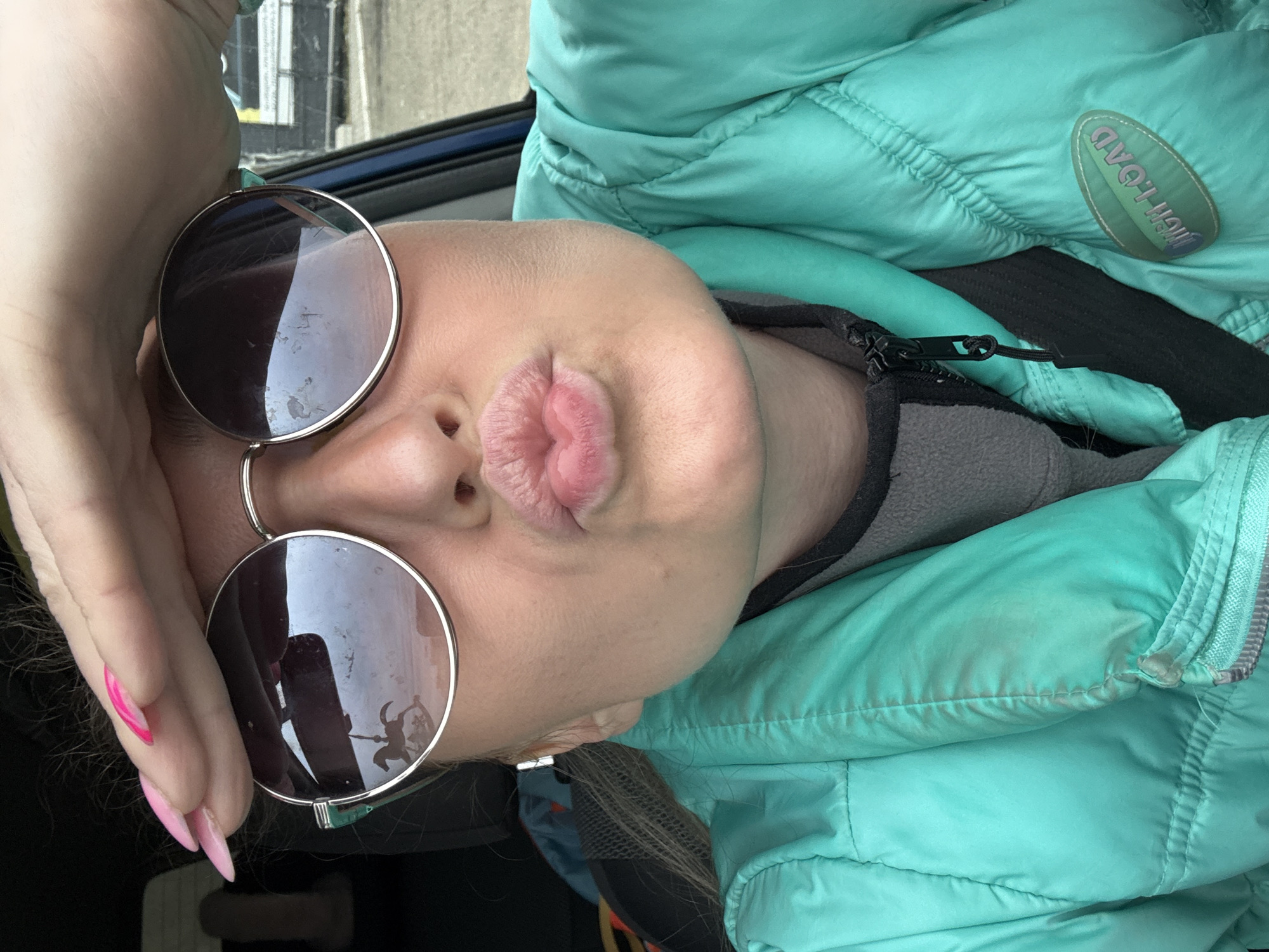 A woman wearing round, dark-tinted sunglasses and a light teal puffer jacket is taking a selfie indoors. She has light skin and is puckering her lips. Her left hand, with long pink nails, is partially visible near her head and glasses. The reflection in her sunglasses shows a building exterior, possibly in the city of Chaikovsky, Russia.