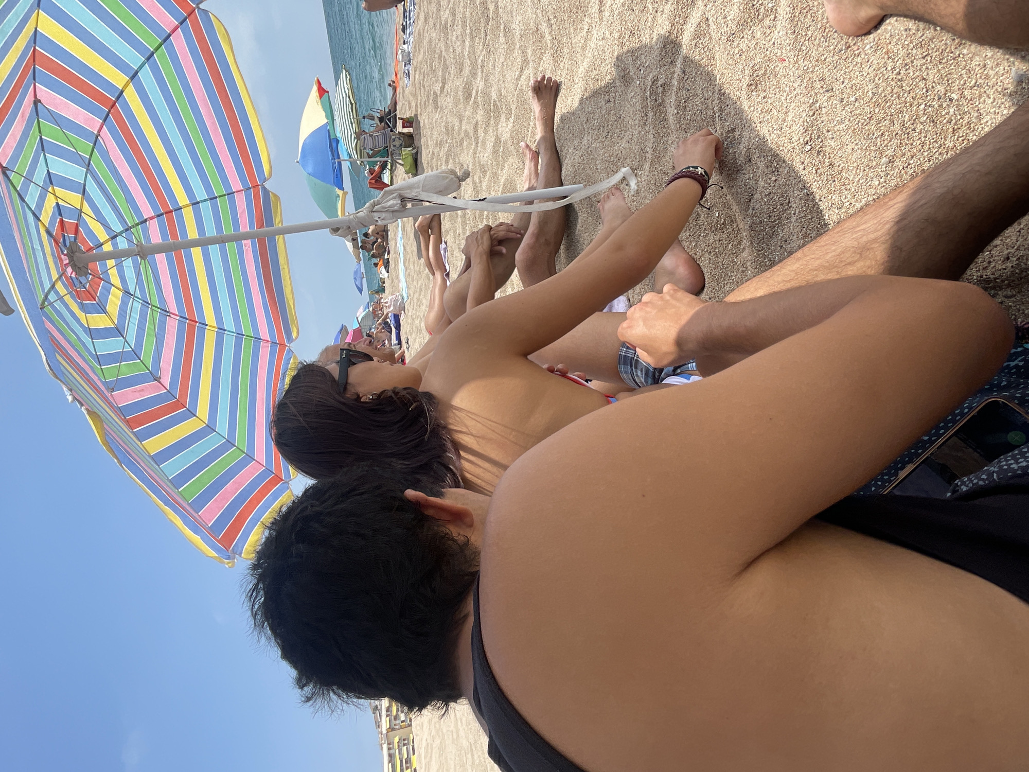 This is a medium shot of people sitting on a beach on a sunny day. In the foreground, a man with dark hair is seen from behind, wearing a black tank top. To his right, a woman with dark hair and sunglasses is seated, facing to the left, wearing a bikini top and checkered shorts. A large, colorful striped beach umbrella shades them. In the background, other people are visible sitting on the sand, some under umbrellas, near the ocean. The scene appears to be on the beach in Canet de Mar, Spain, given the presence of beach umbrellas and the general atmosphere of a coastal town. The sand is light brown and textured, and the water is a calm blue.