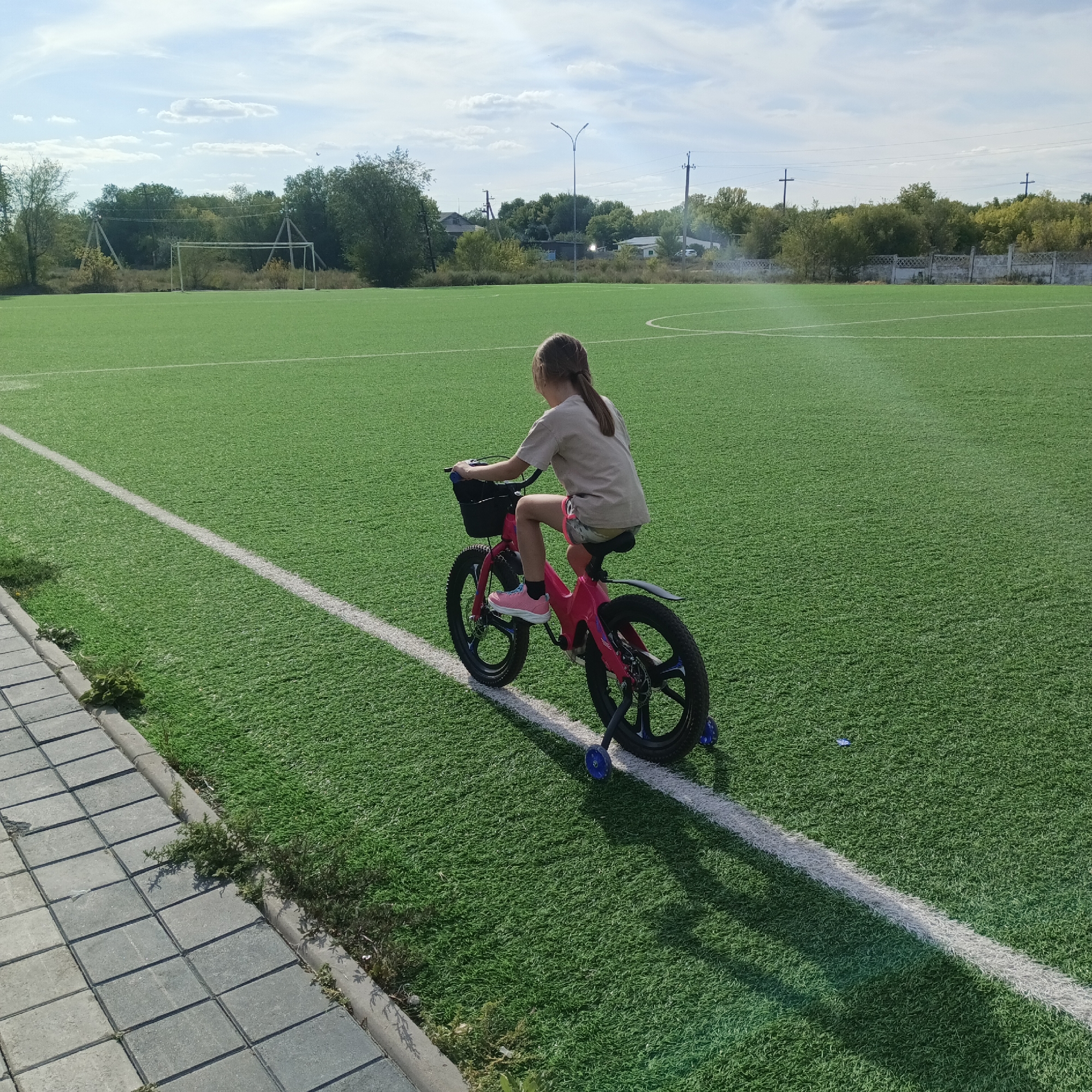 A young girl with brown hair in a ponytail, wearing a light brown t-shirt and patterned shorts, rides a pink bicycle on a green artificial turf field. The bicycle has training wheels and a black basket on the handlebars. A white line marks the edge of a sports field, with a paved walkway to the left of the field. In the background, trees, utility poles, a soccer goal, and some buildings are visible under a bright blue sky with scattered clouds. The scene appears to be outdoors on a sunny day.